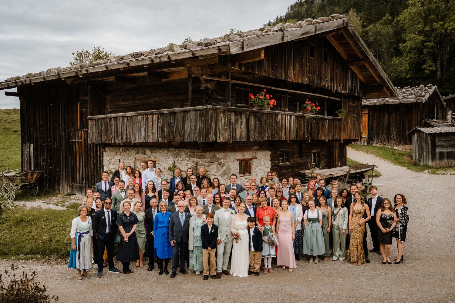 Large wedding party group photo taken outside rustic barn with mountain landscape in background.