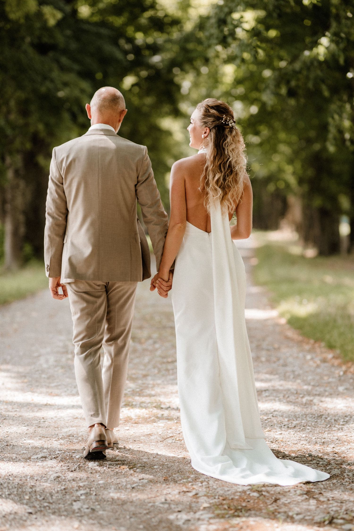 A couple in wedding attire walks hand in hand down a tree-lined gravel path on their special day.