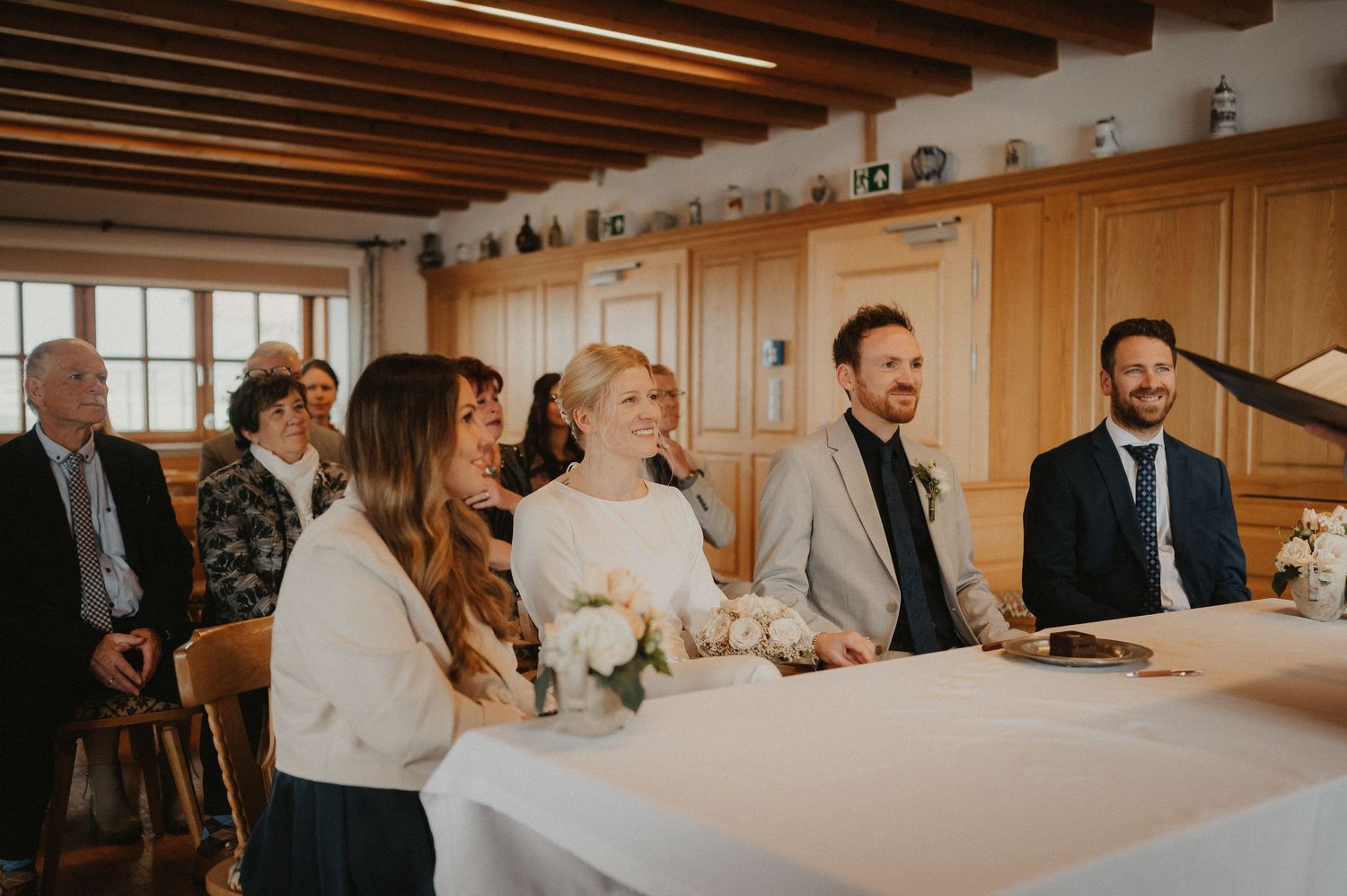 Wedding reception scene with guests seated at tables in a warm-lit room with wooden paneled walls.