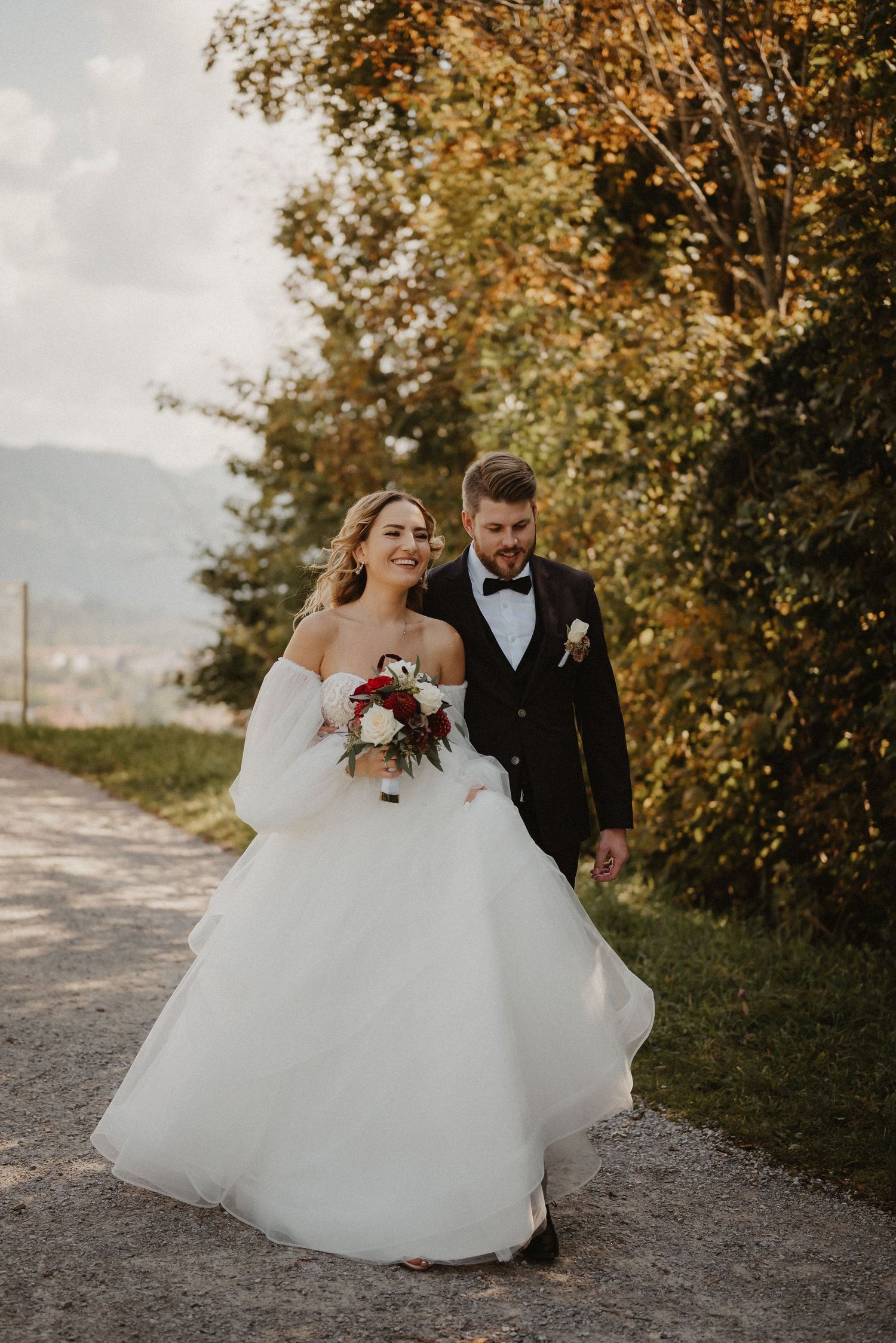 Newlyweds posing on a dirt path surrounded by autumn trees and mountain views.