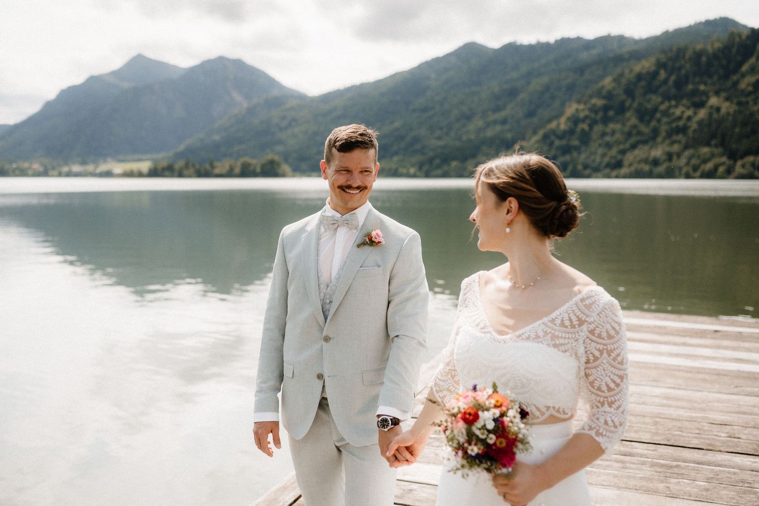 A couple in wedding attire walks along a wooden pier with scenic mountain views.