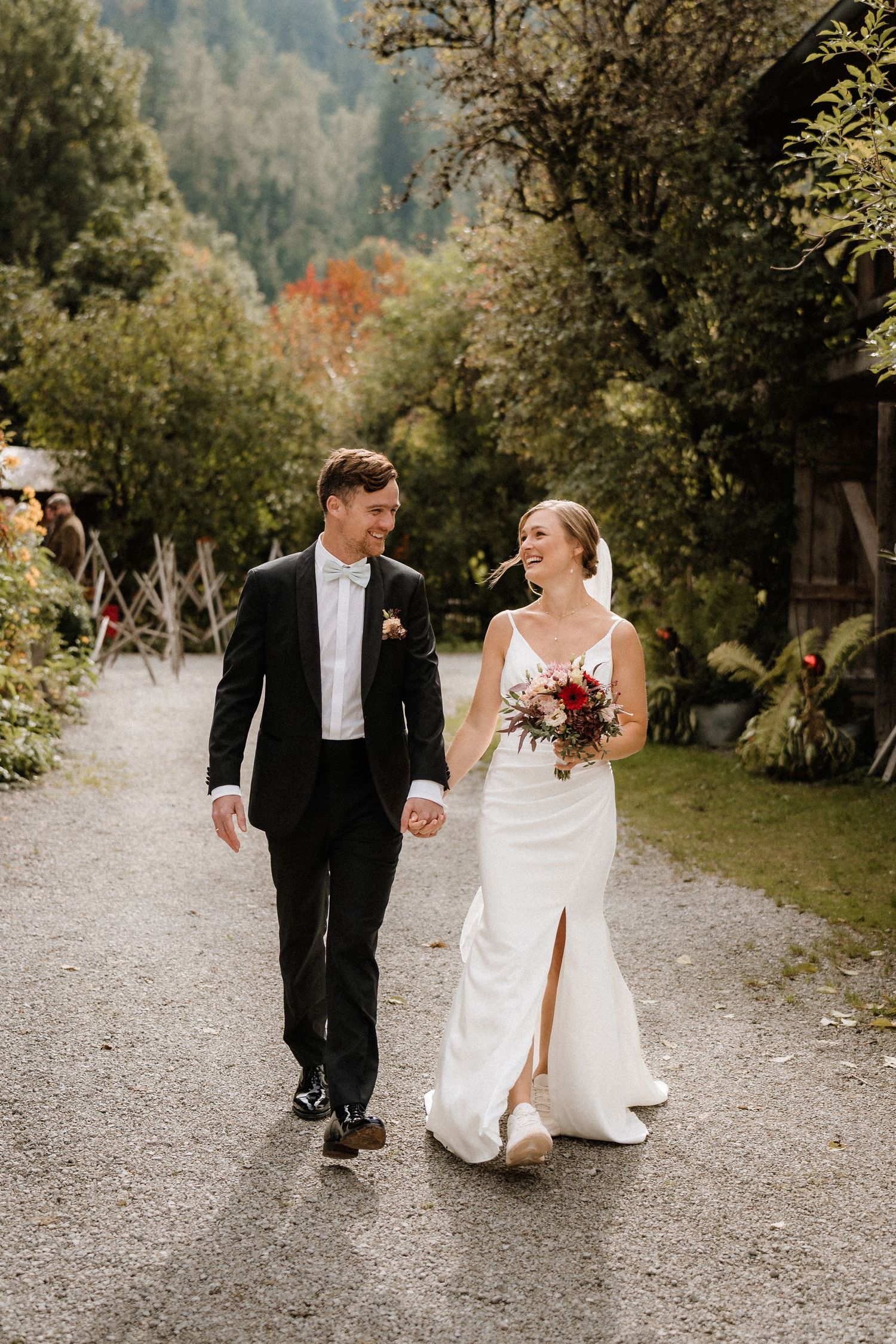 Hochzeitspaar geht zusammen auf einem Kiesweg, umgeben von Herbstbäumen. Von Dama Fotografie Bad Tölz