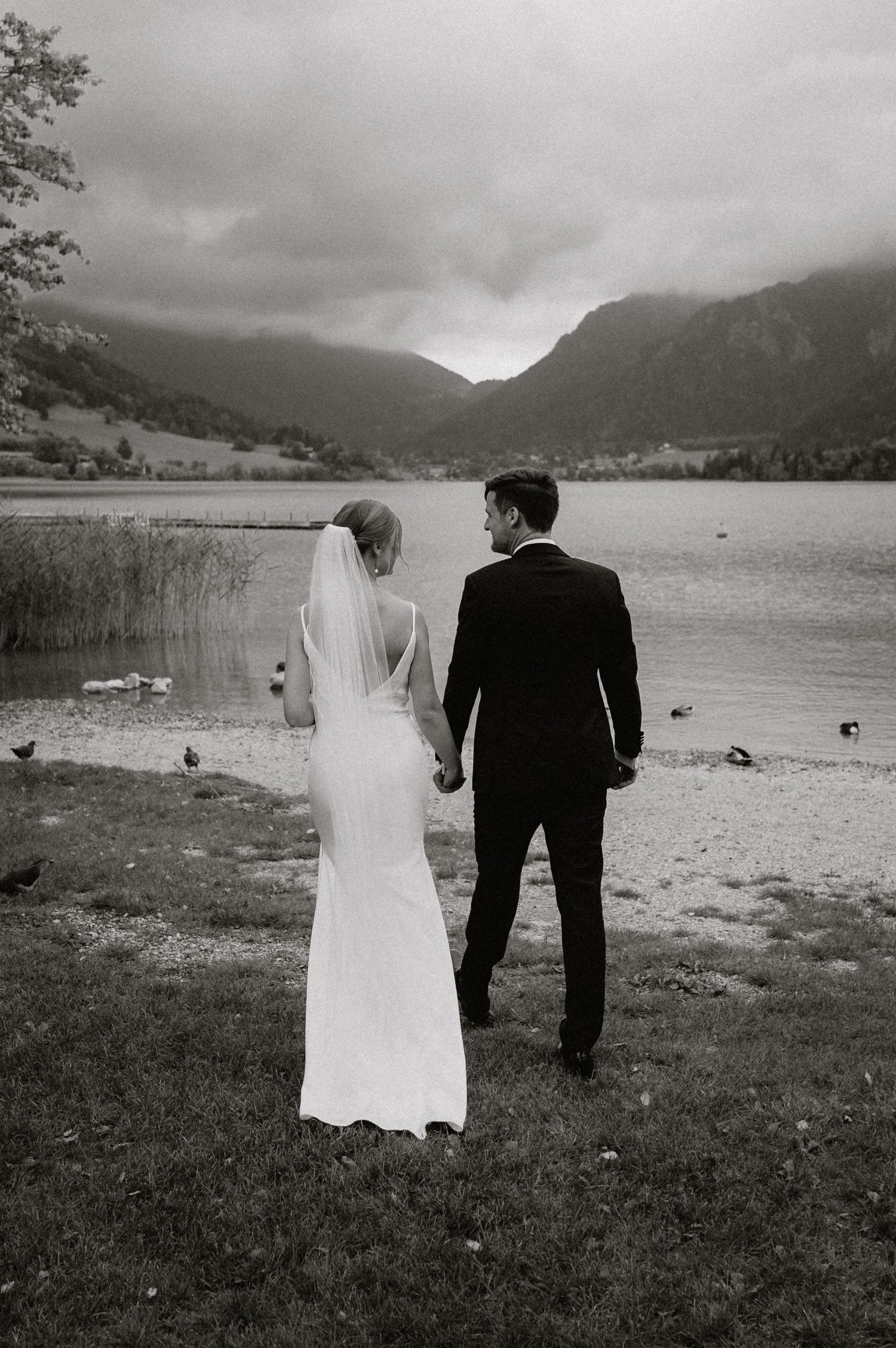 Couple in wedding attire stands by lakeside with misty mountains in background.