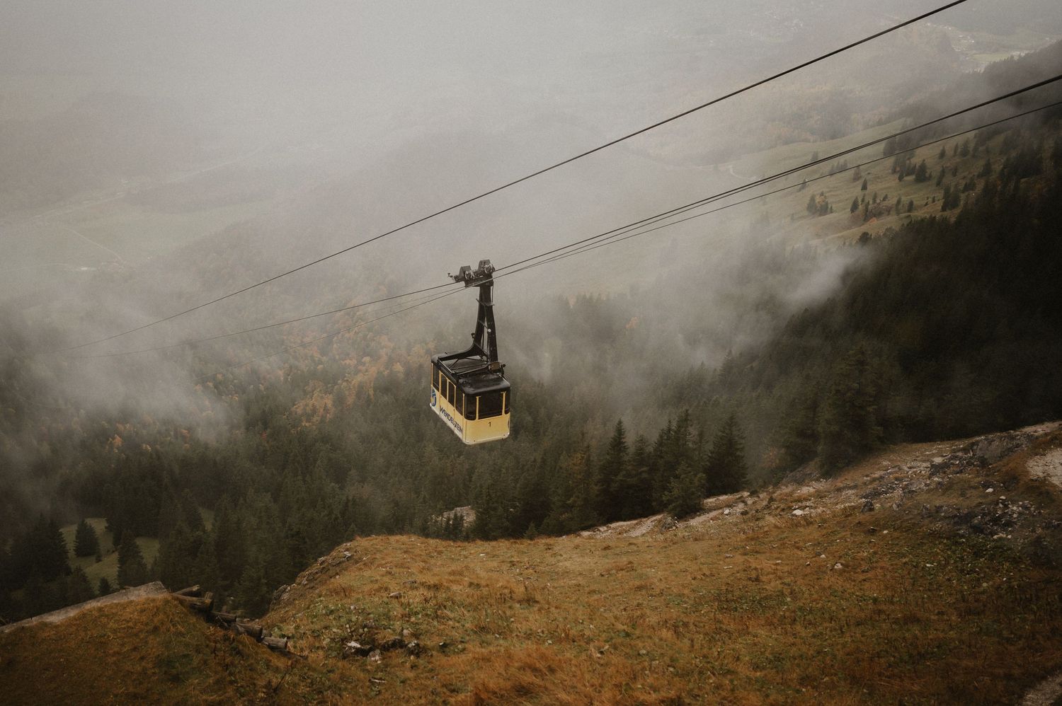 Cable car gondola moving through foggy mountain terrain with power lines visible.