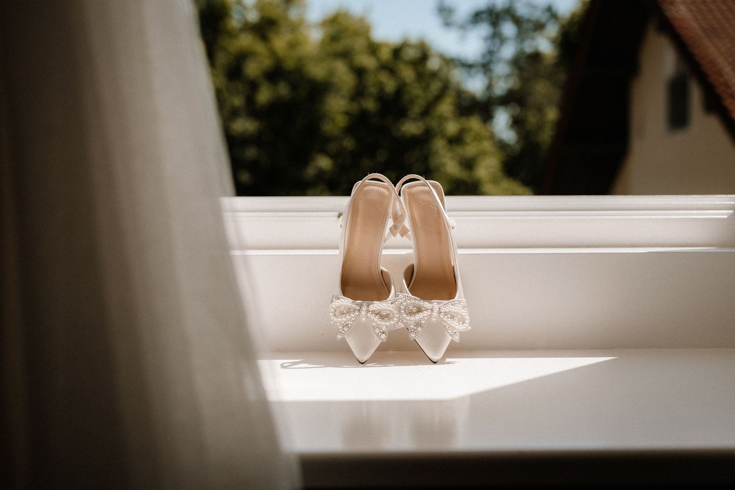 Elegant bridal shoes sit on a white windowsill in natural sunlight.