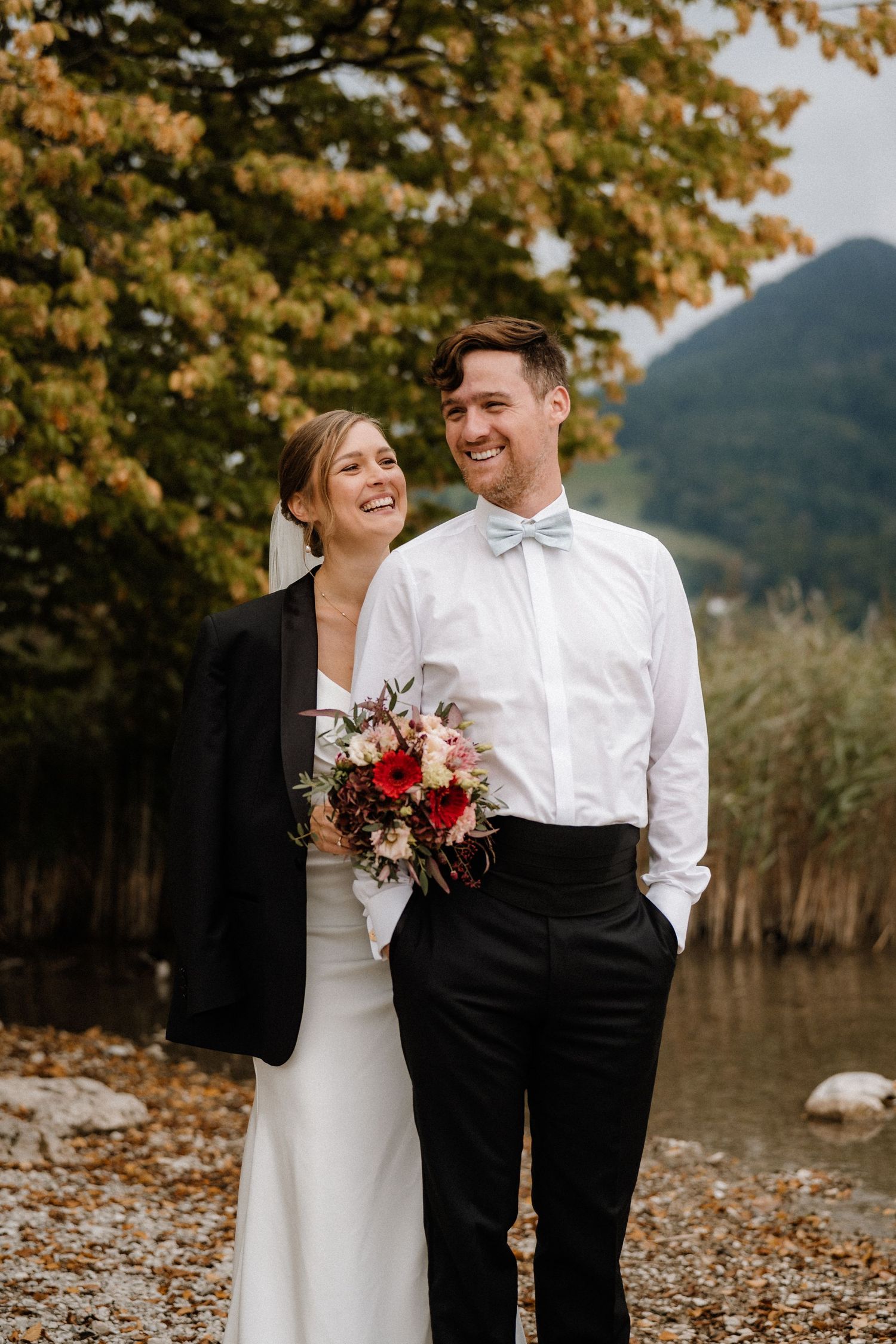 Happy couple in wedding attire laughing together by a lakeside with autumn trees.
