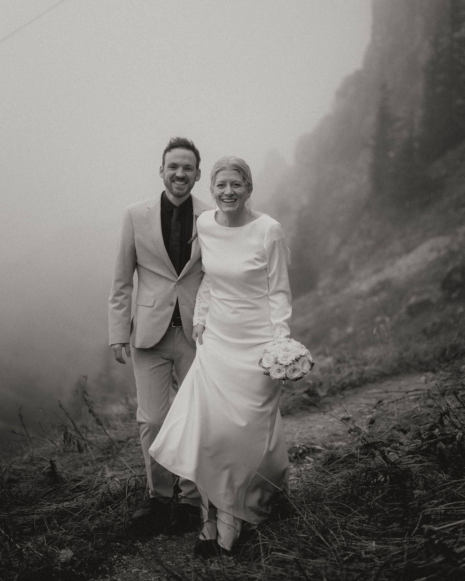 Black and white photo of newlyweds walking along a misty mountain path.