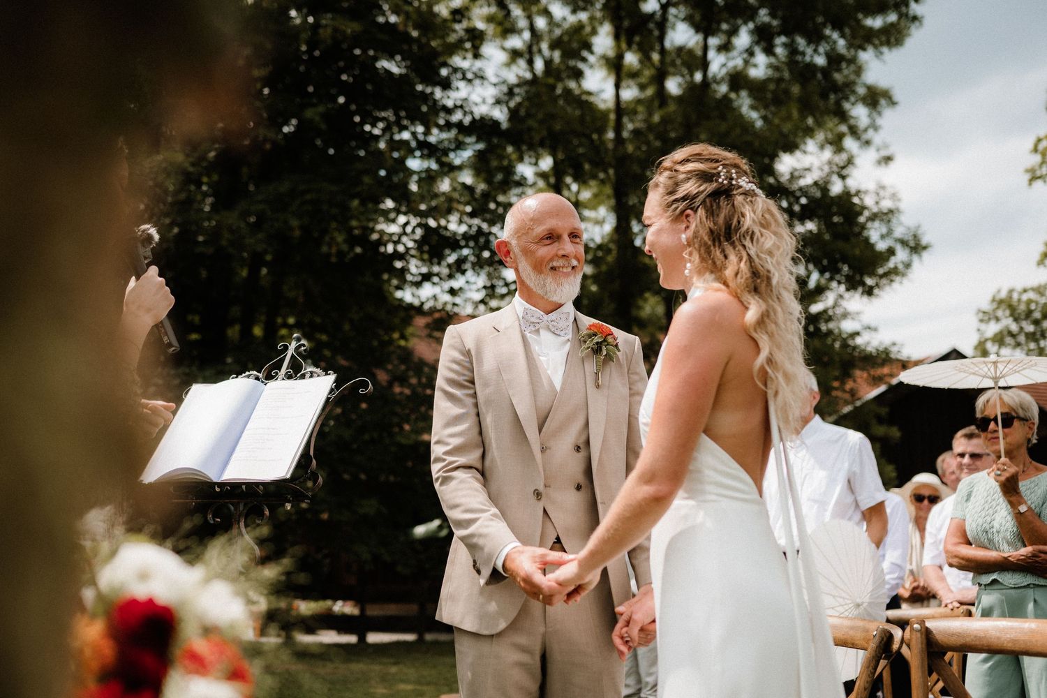 Outdoor wedding ceremony with guests seated in wooden chairs and floral arrangements.