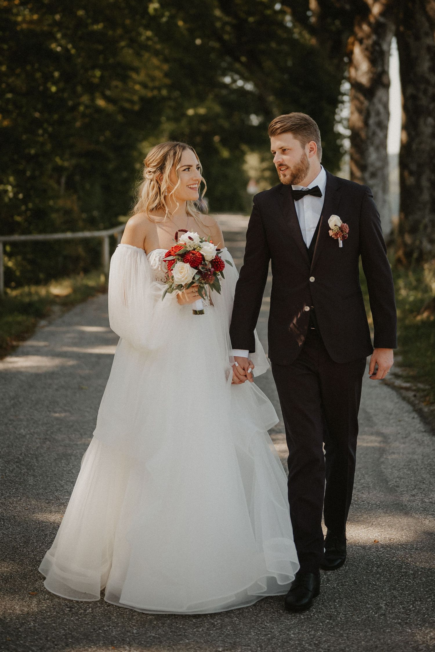 A couple walks hand in hand on a path, with the bride in a white gown and groom in classic tuxedo.