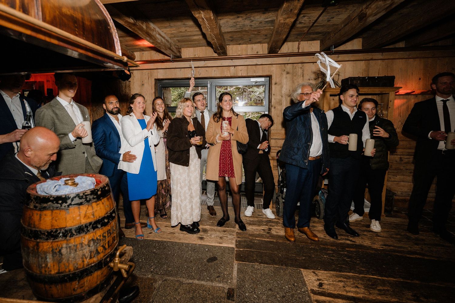Wedding celebration in a rustic barn with guests gathered around wooden barrels.