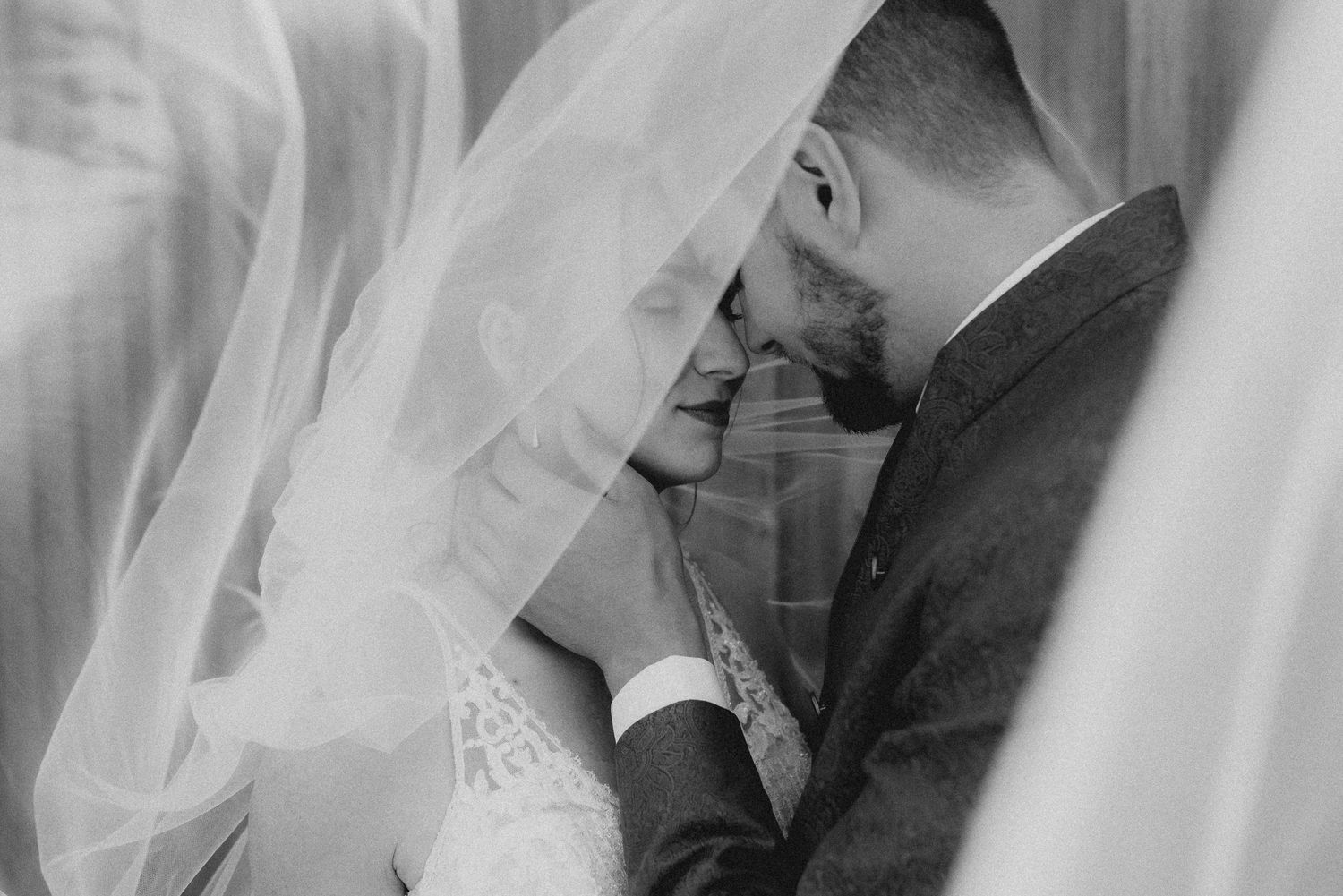 Ein romantisches Schwarz-Weiß-Hochzeitsfoto zeigt ein Paar, das einen intimen Moment hinter einem fließenden Schleier teilt. Von Dama Foto
