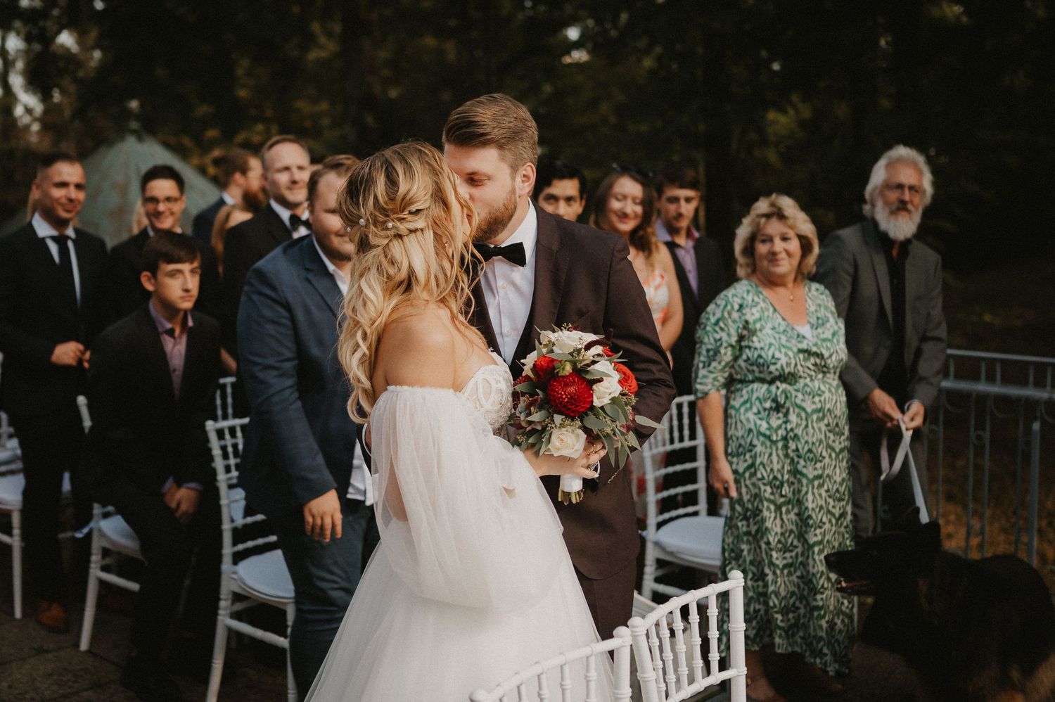 Outdoor wedding ceremony setup with white chairs and guests gathered on a sunny afternoon.