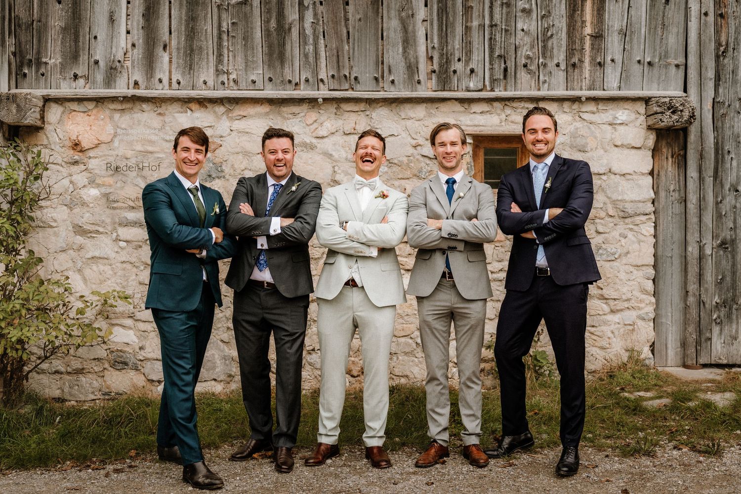 Groomsmen posing against weathered wooden barn wall in formal attire.
