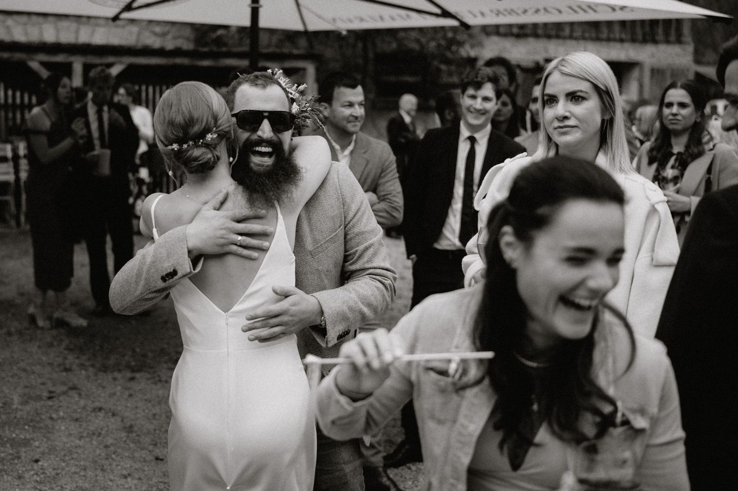 Wedding party celebrates outdoors with umbrellas near wooden barn structures.