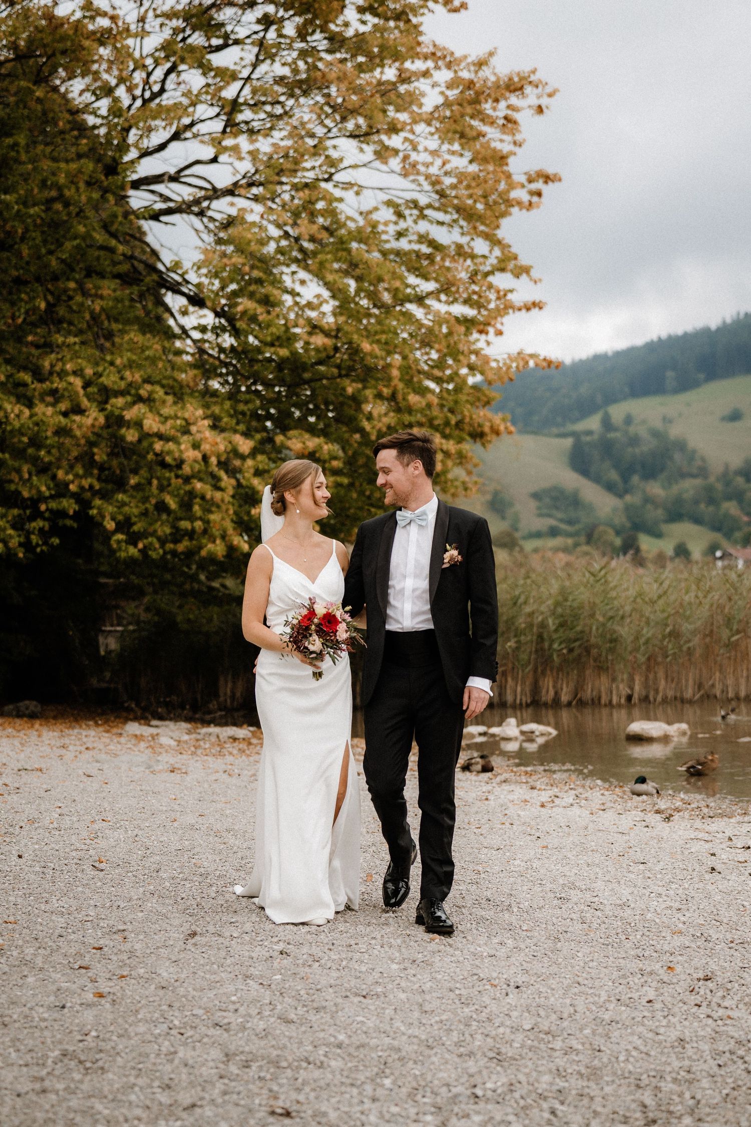 Couple walks along lakeside path with autumn foliage and mountains in background.
