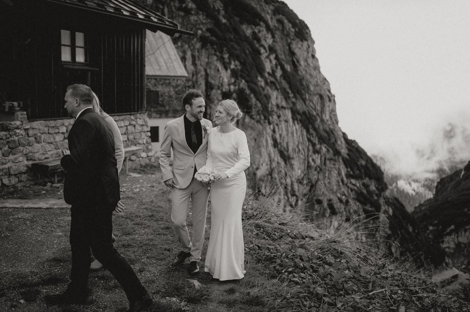 A wedding celebration takes place on a mountainside path next to a stone building with dramatic cliffs in the background.