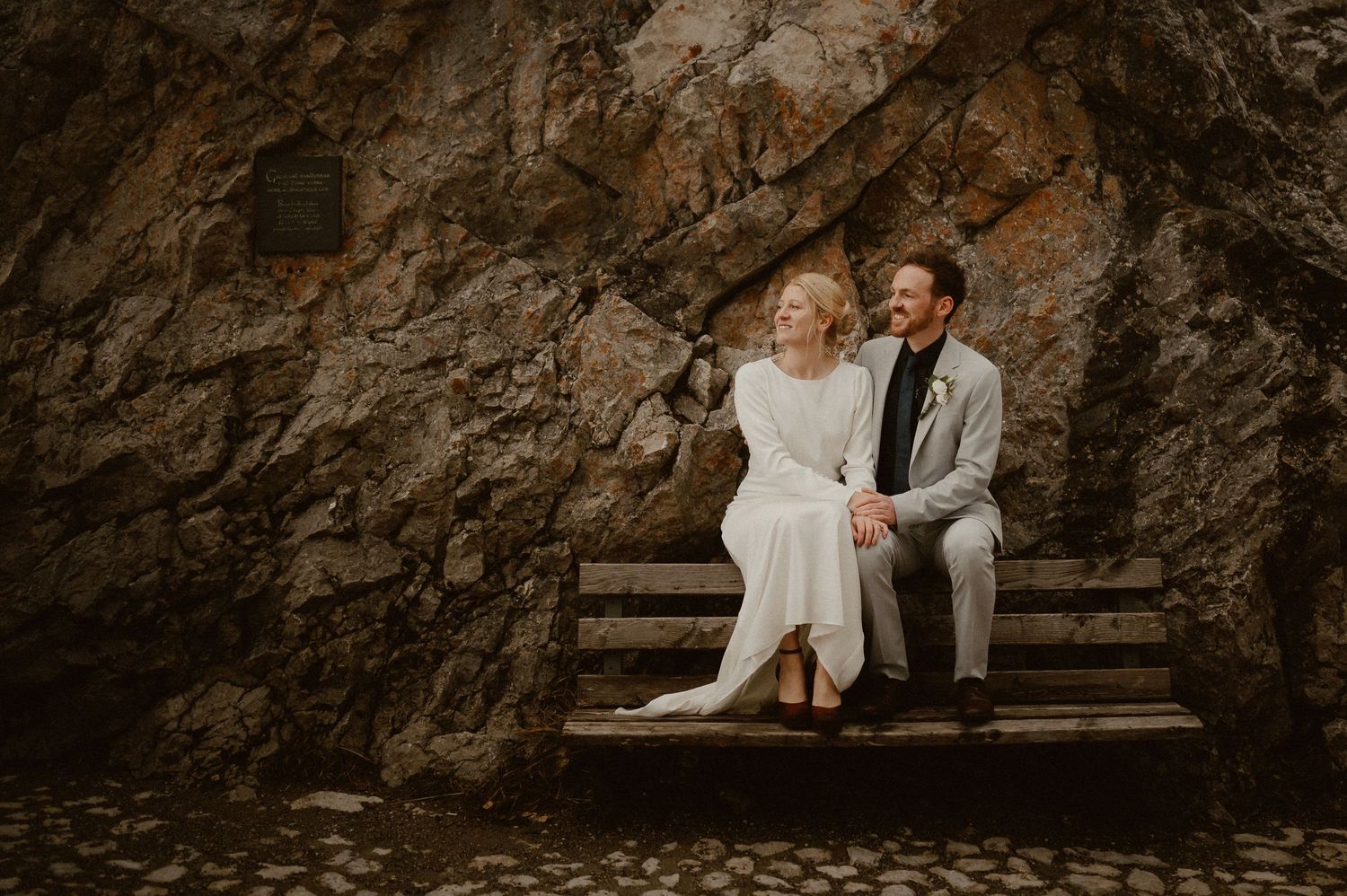 Two people sit together on stone steps against a dramatic rocky cliff backdrop.