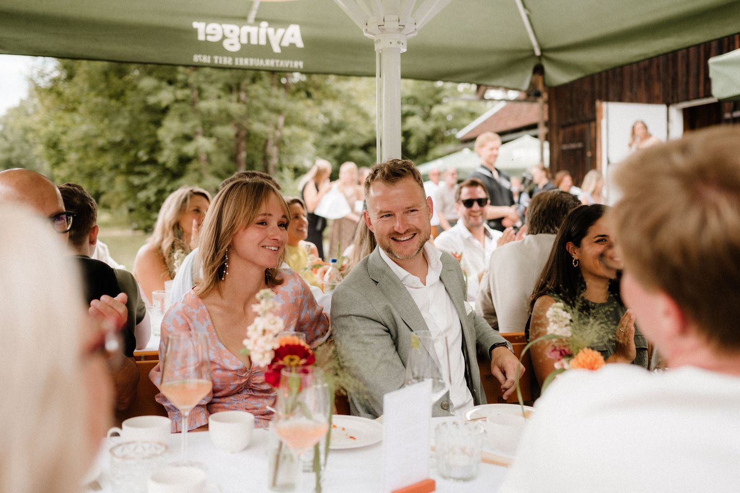 Outdoor reception scene with guests seated under white tent canopy.
