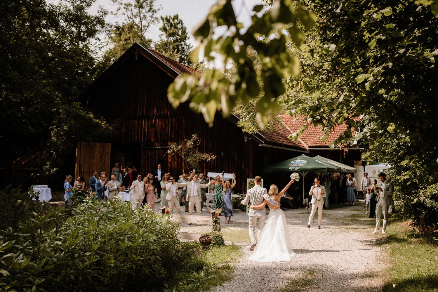 Weitwinkelansicht der Hochzeitsfeier mit Gästen, die sich an einem rustikalen Veranstaltungsort im Freien versammelt haben. Von Dama Foto