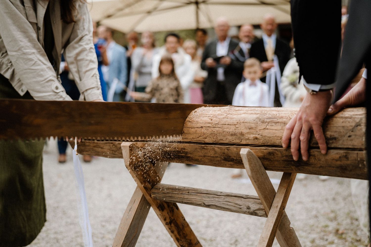 Wedding party gathers around wooden log for traditional German sawing ceremony.