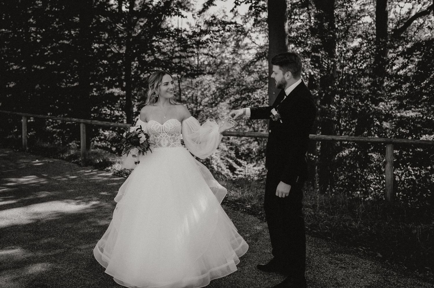 A black and white wedding photo of a couple in formal attire standing in a wooded setting.