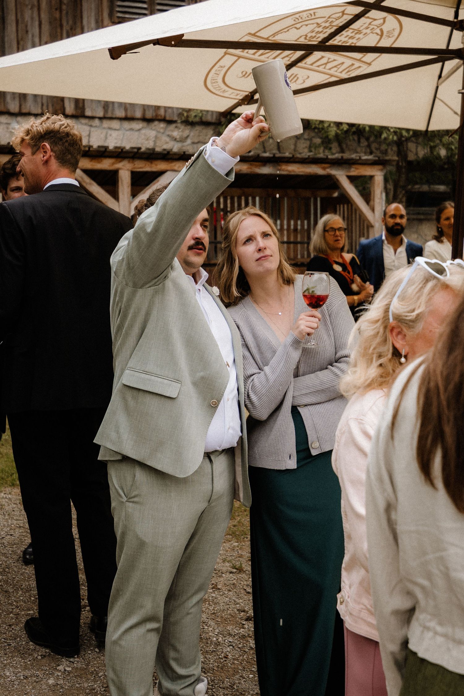 Wedding guest raises glass during outdoor reception while others mingle beneath umbrella.