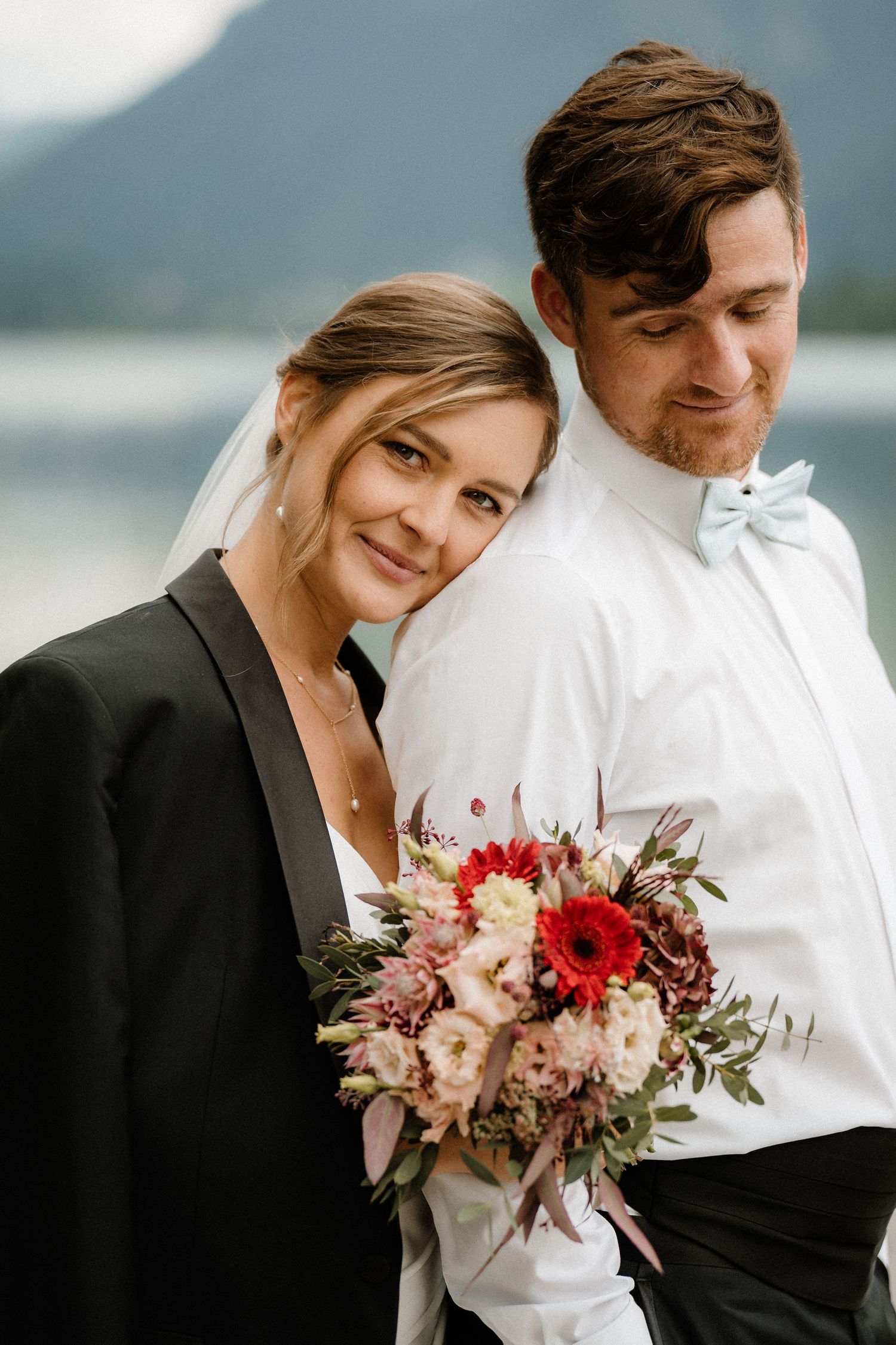 Couple in formal attire sharing an intimate moment with red and pink floral bouquet.
