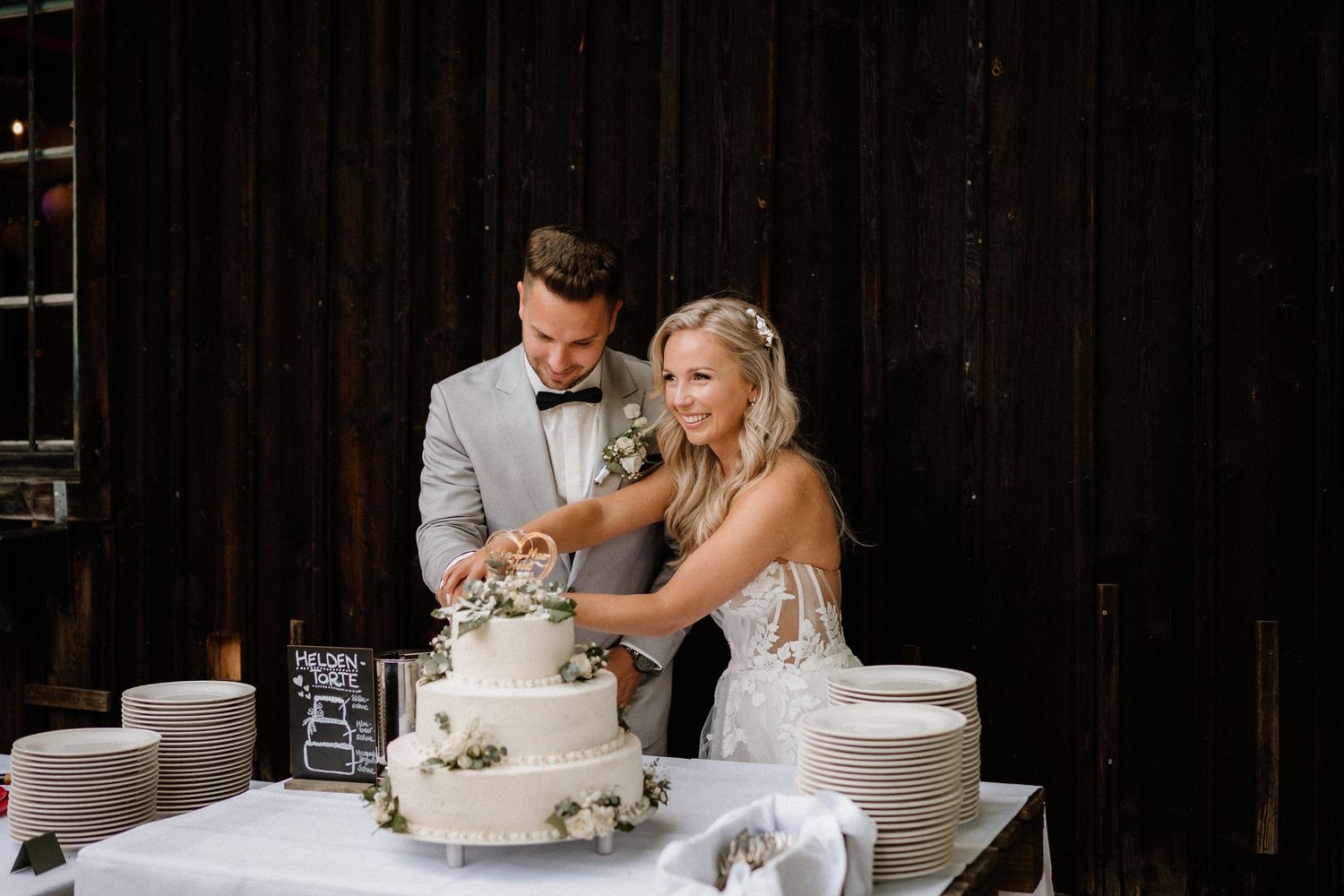 Newlyweds cutting their elegant white wedding cake together in a rustic barn setting.