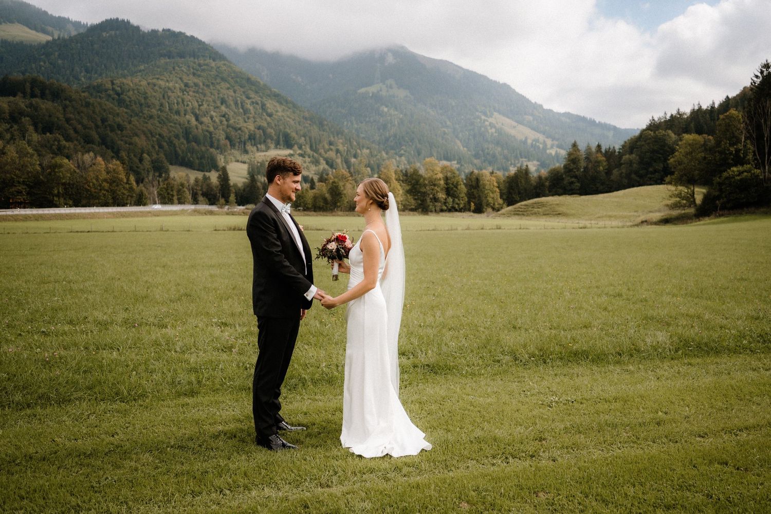 Wedding couple stands in meadow with majestic mountain backdrop on sunny day.