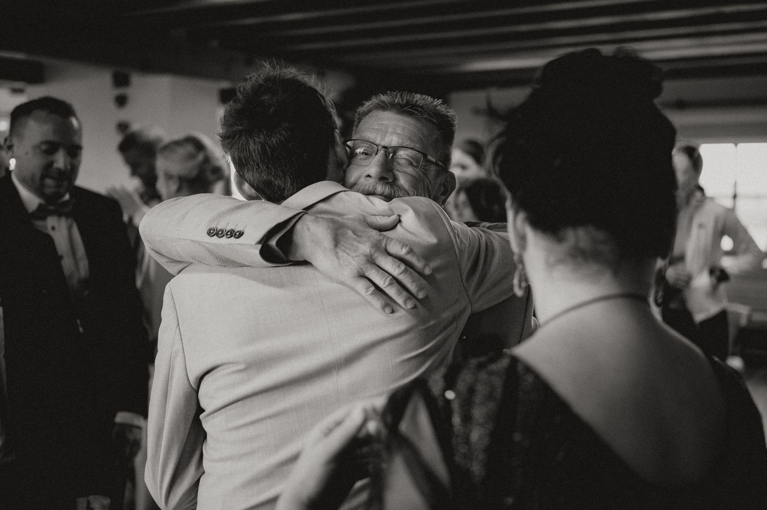 Intimate black and white photograph of wedding guests embracing during reception celebrations.
