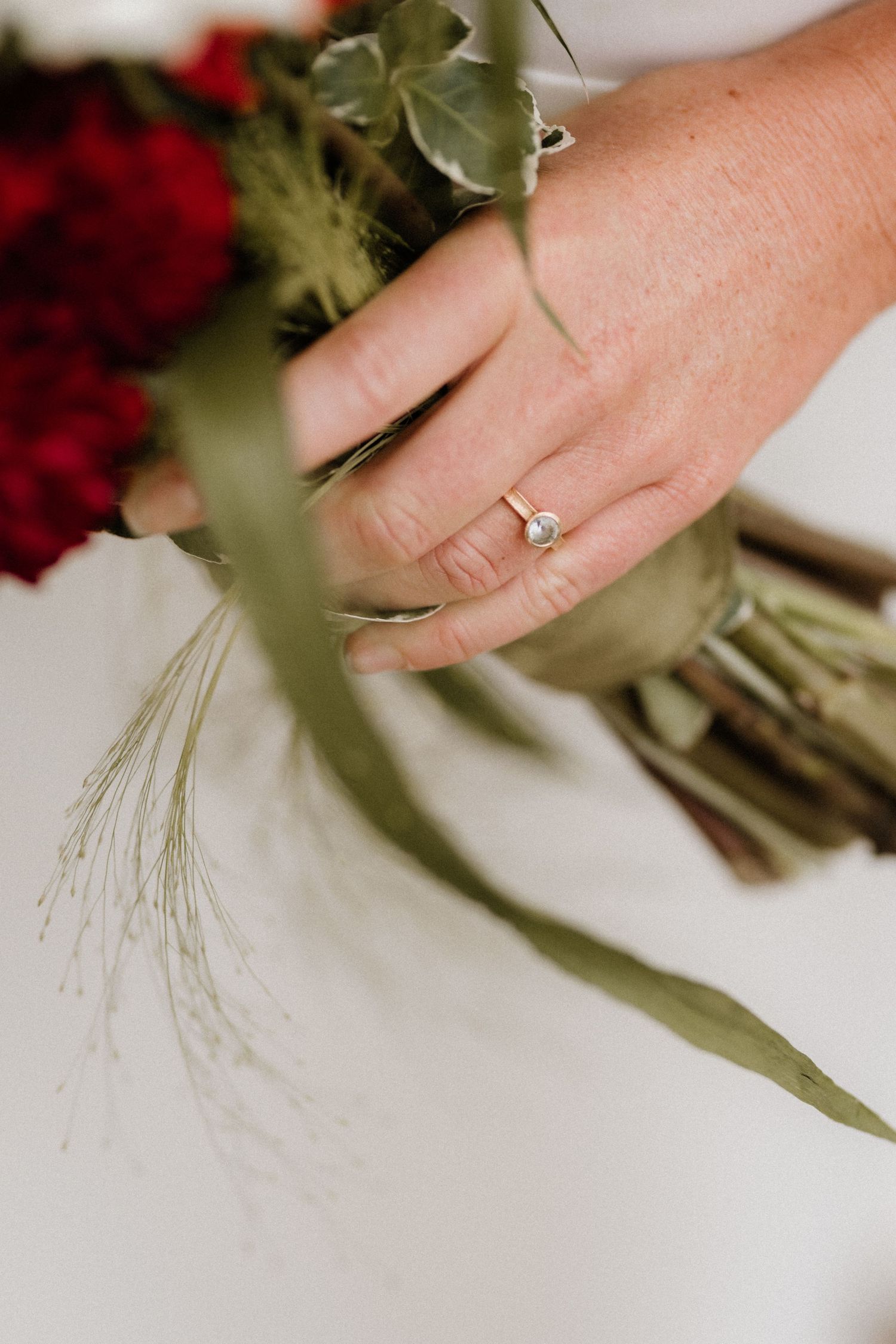 Close-up of an engagement ring on a hand holding a bouquet with red flowers and greenery.