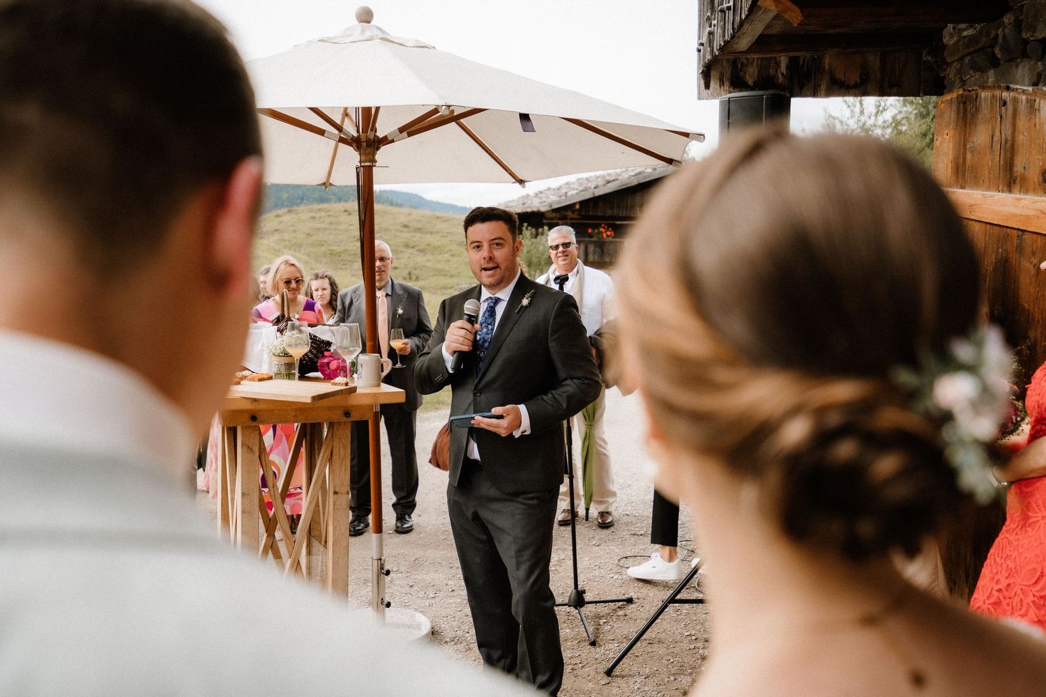 Outdoor wedding celebration with guests gathered under white umbrellas near rustic buildings.