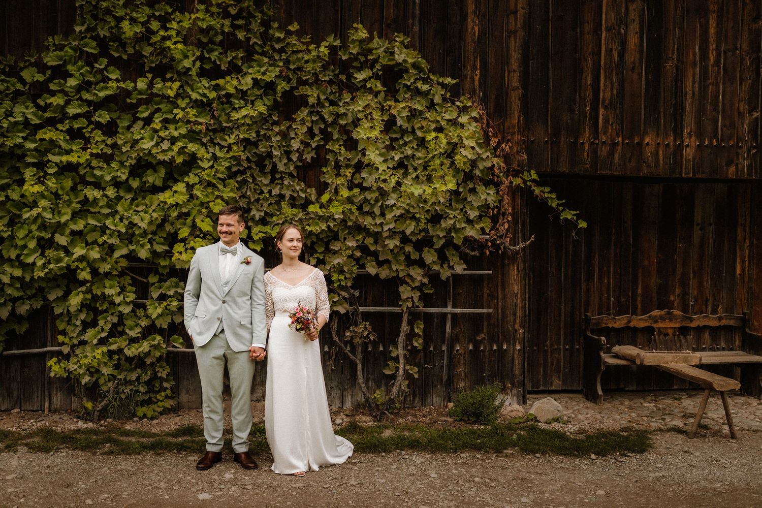 Wedding couple poses in front of vine-covered rustic wooden barn wall.