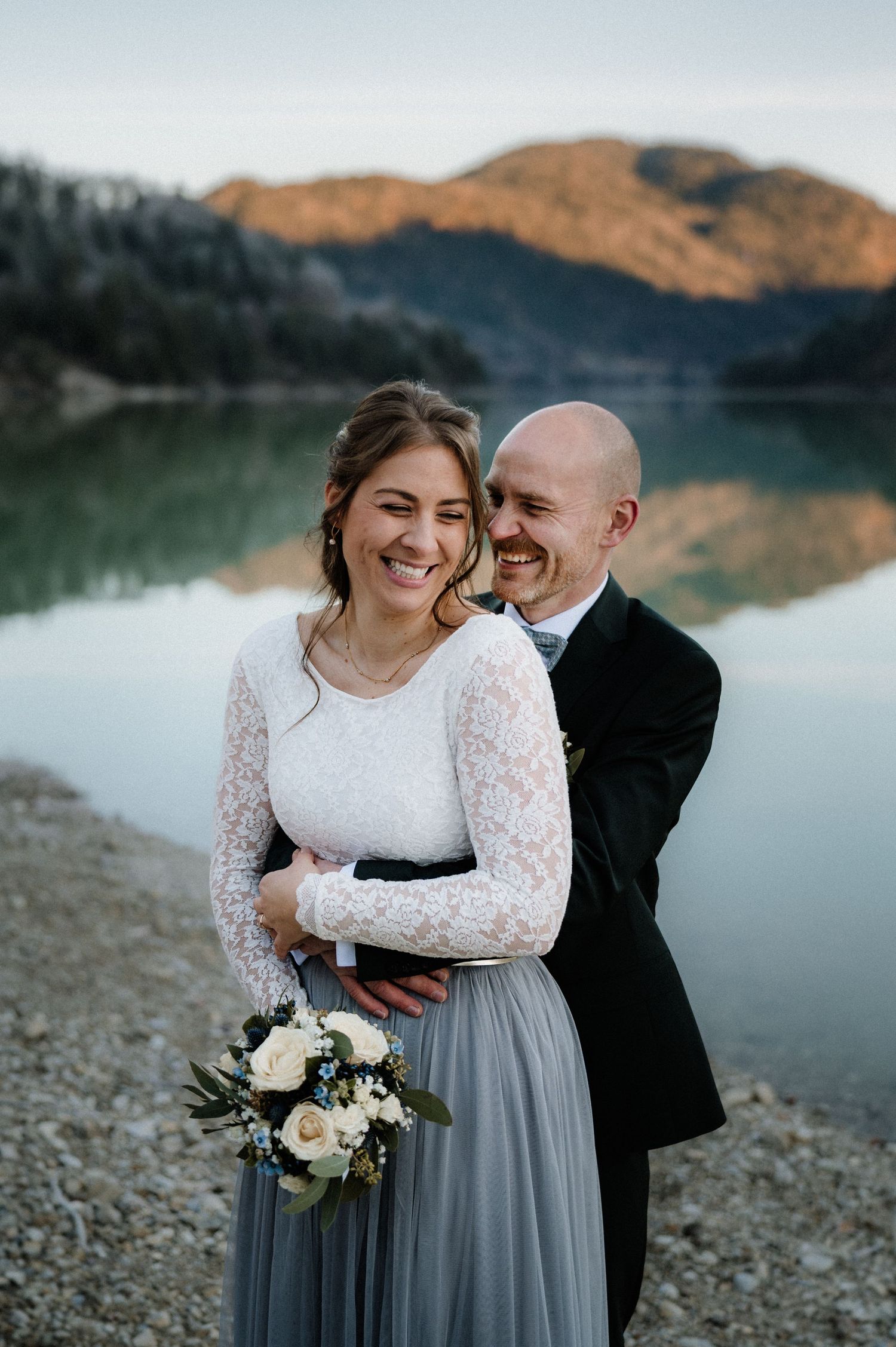 Ein Paar teilt einen freudigen Moment an einem ruhigen Bergsee, wobei die Braut ein Spitzenoberteil und einen grauen Rock trägt und Blumen.