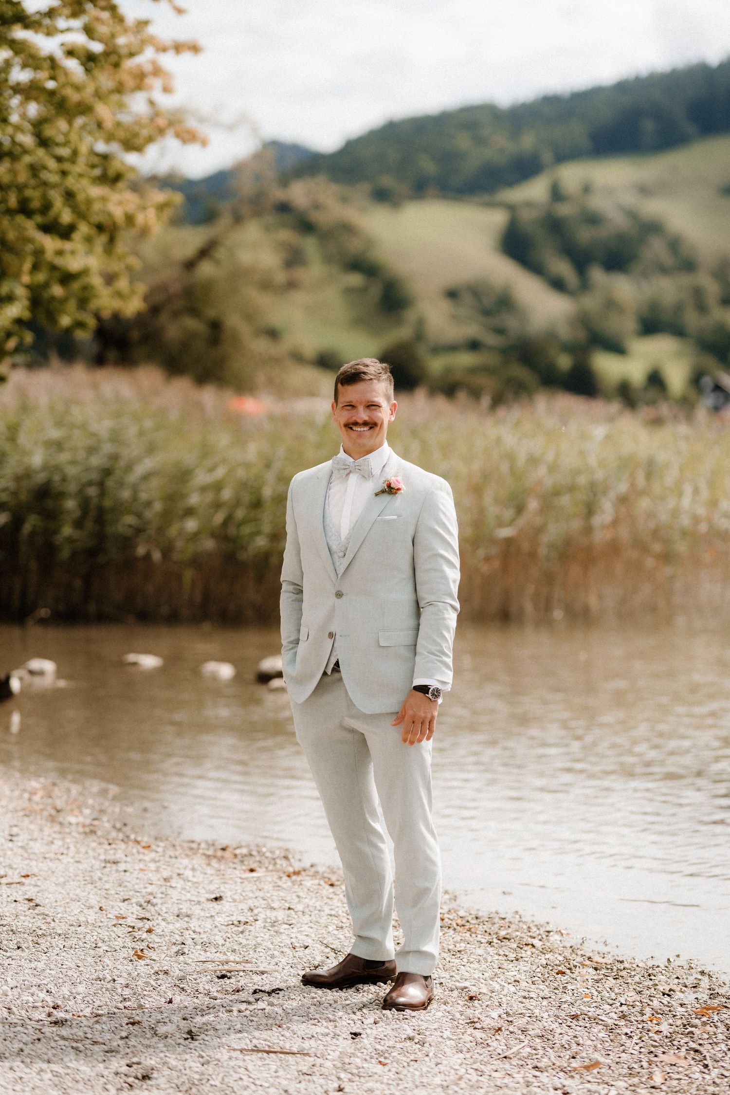 A groom in a light gray suit stands by a lakeside with mountains in the background.
