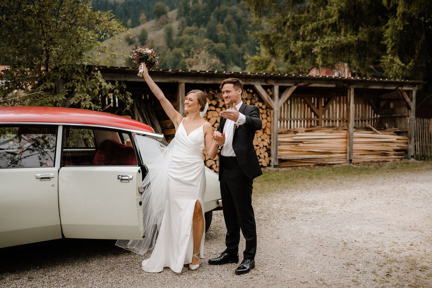 Wedding party gathered around vintage cars on rustic gravel driveway.