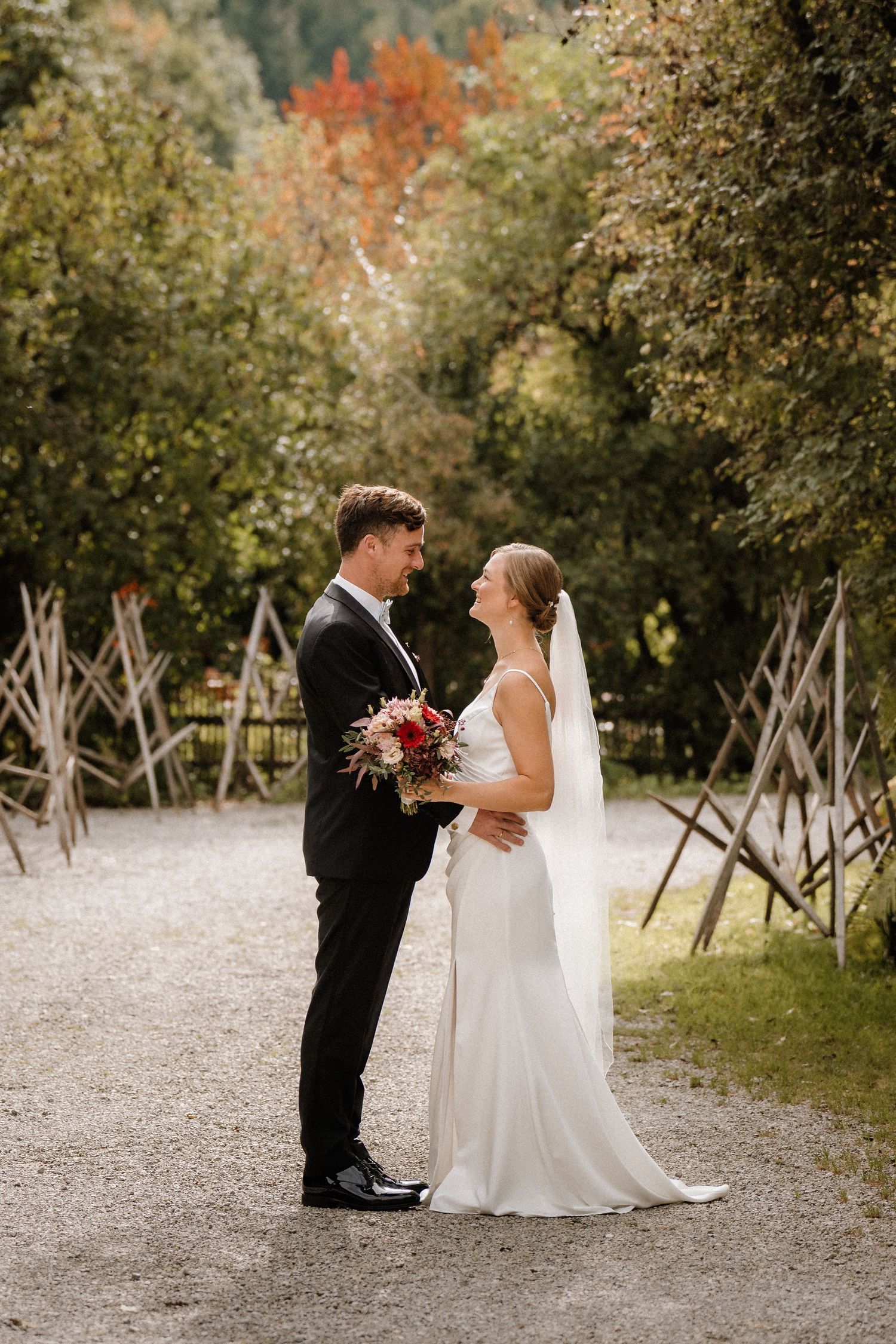 Wedding couple on gravel path surrounded by autumn foliage and wooden trellises.