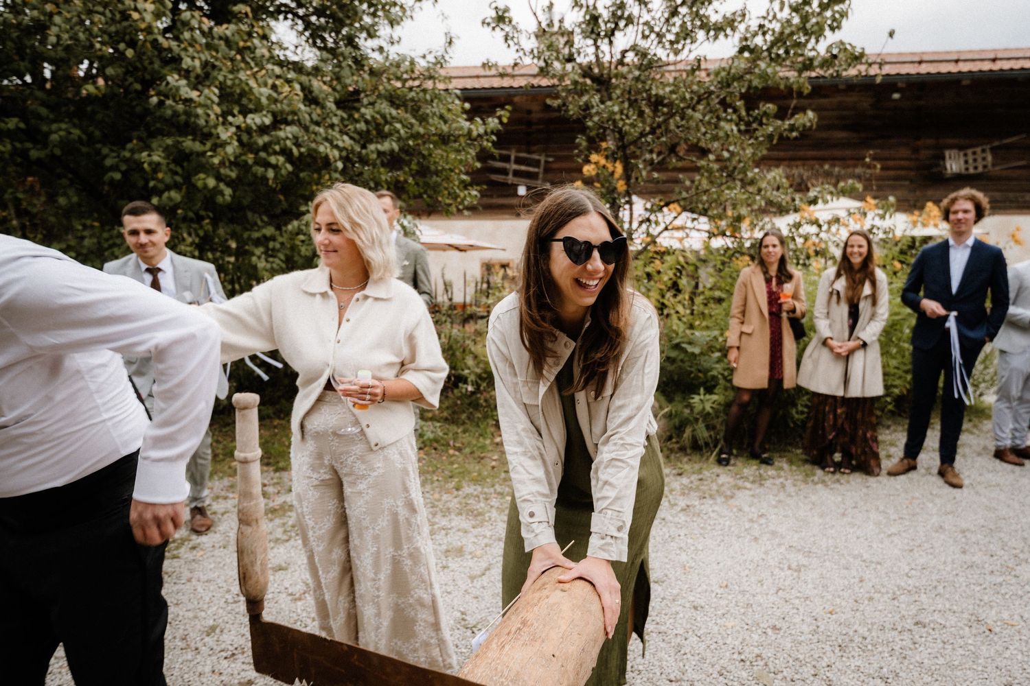 Wedding guests participate in a traditional log sawing activity outside a rustic venue.