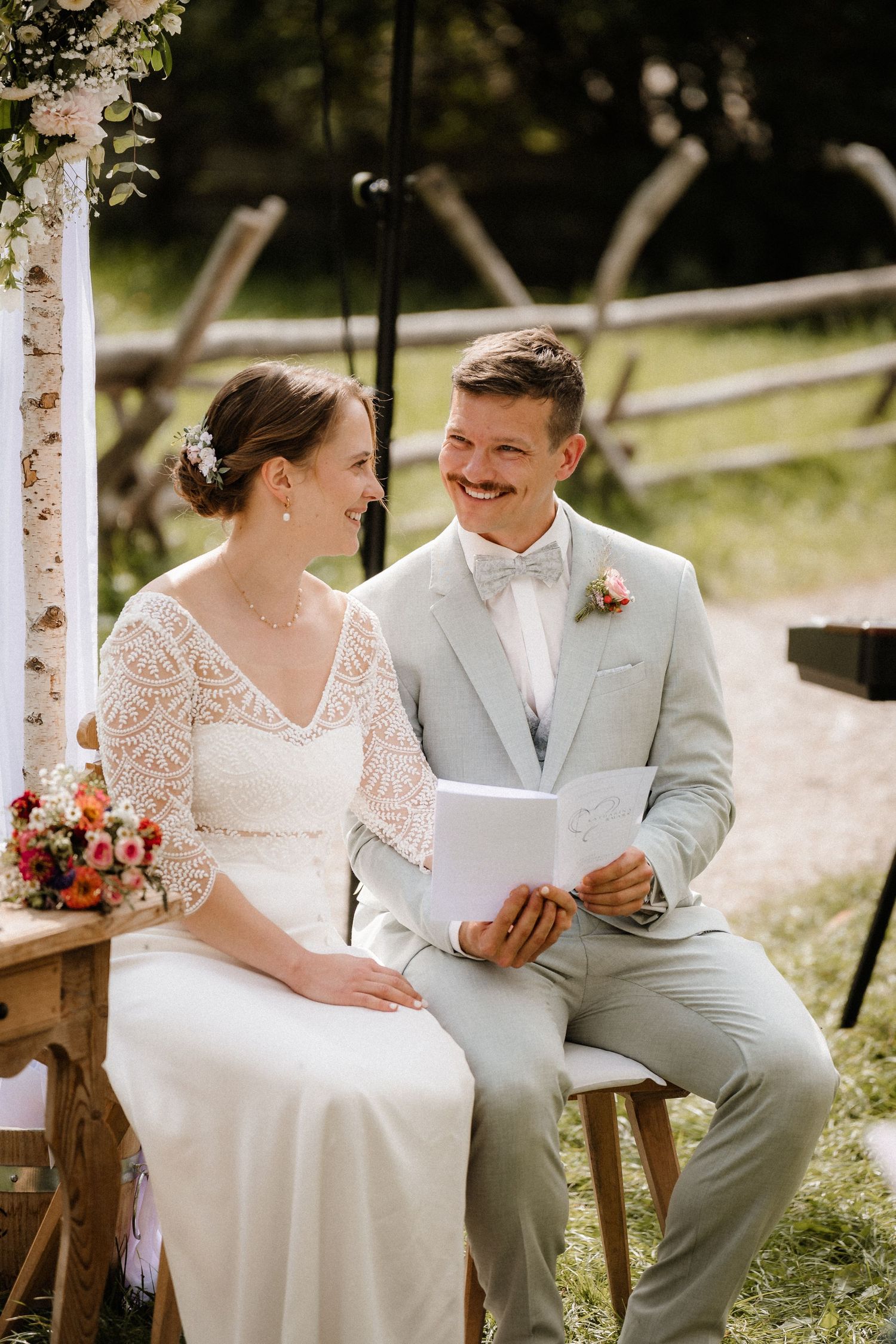 Outdoor garden wedding ceremony setup with couple seated on wooden chairs under floral decor.