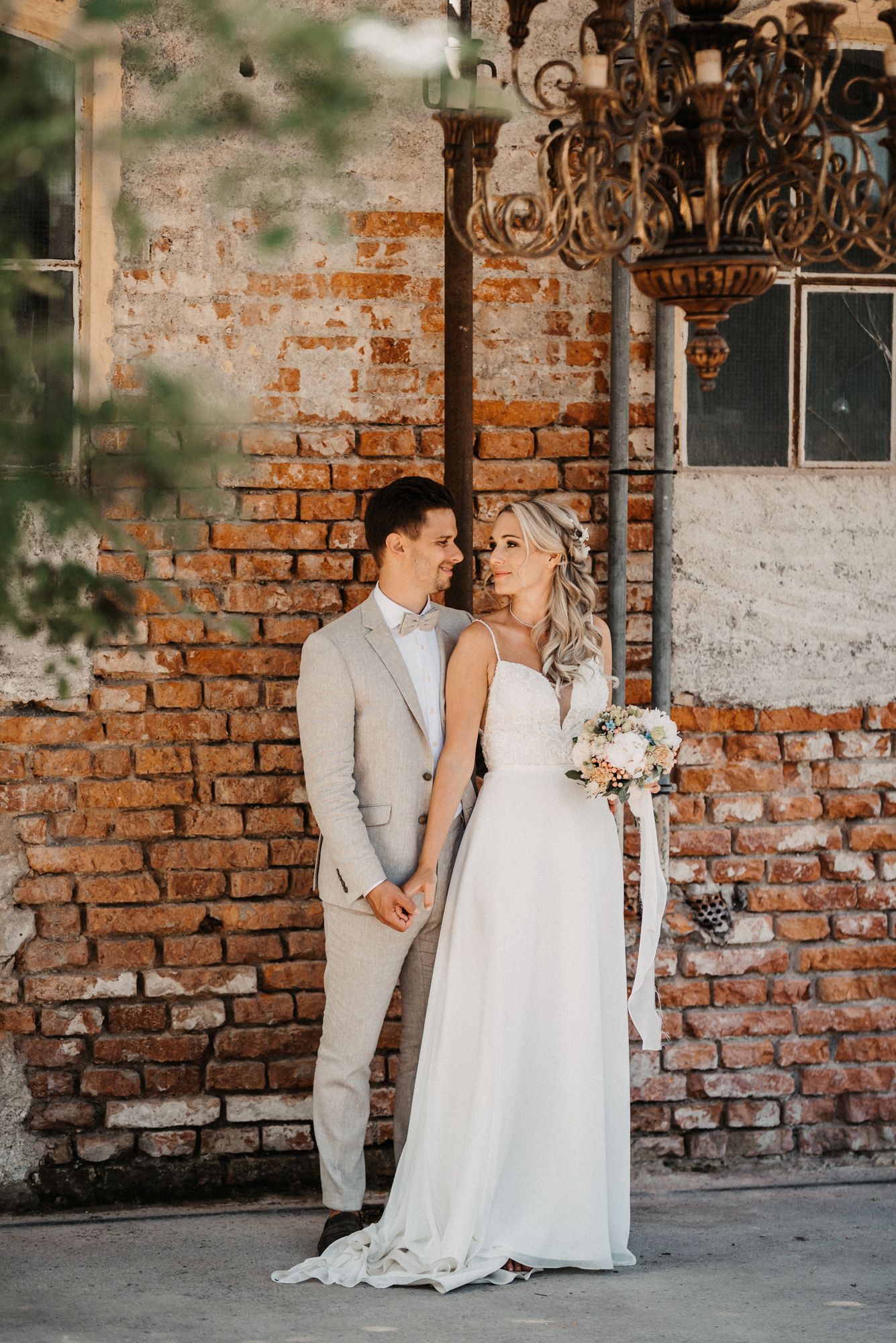 Ein romantisches Hochzeitspaar posiert vor einer freiliegenden Backsteinmauer mit eleganten Kronleuchtern. Von Dama Fotografie Bad Tölz