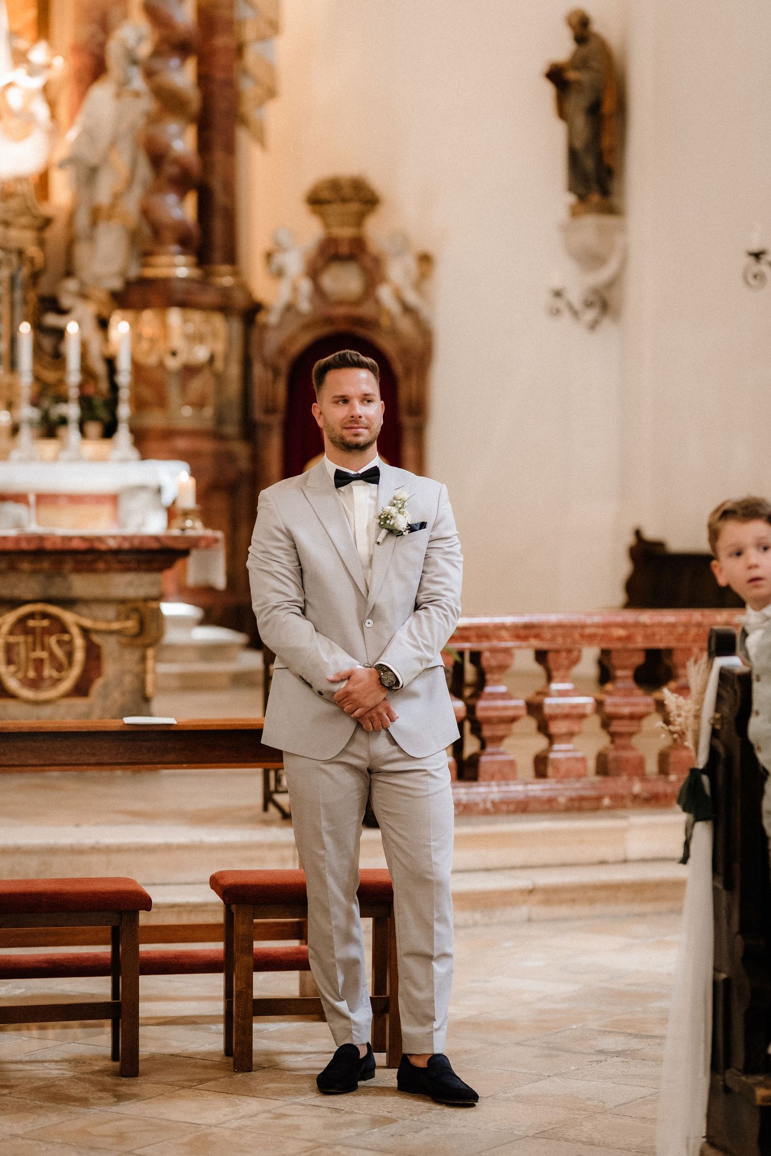 Wedding ceremony in church with groom in light gray suit standing at altar.