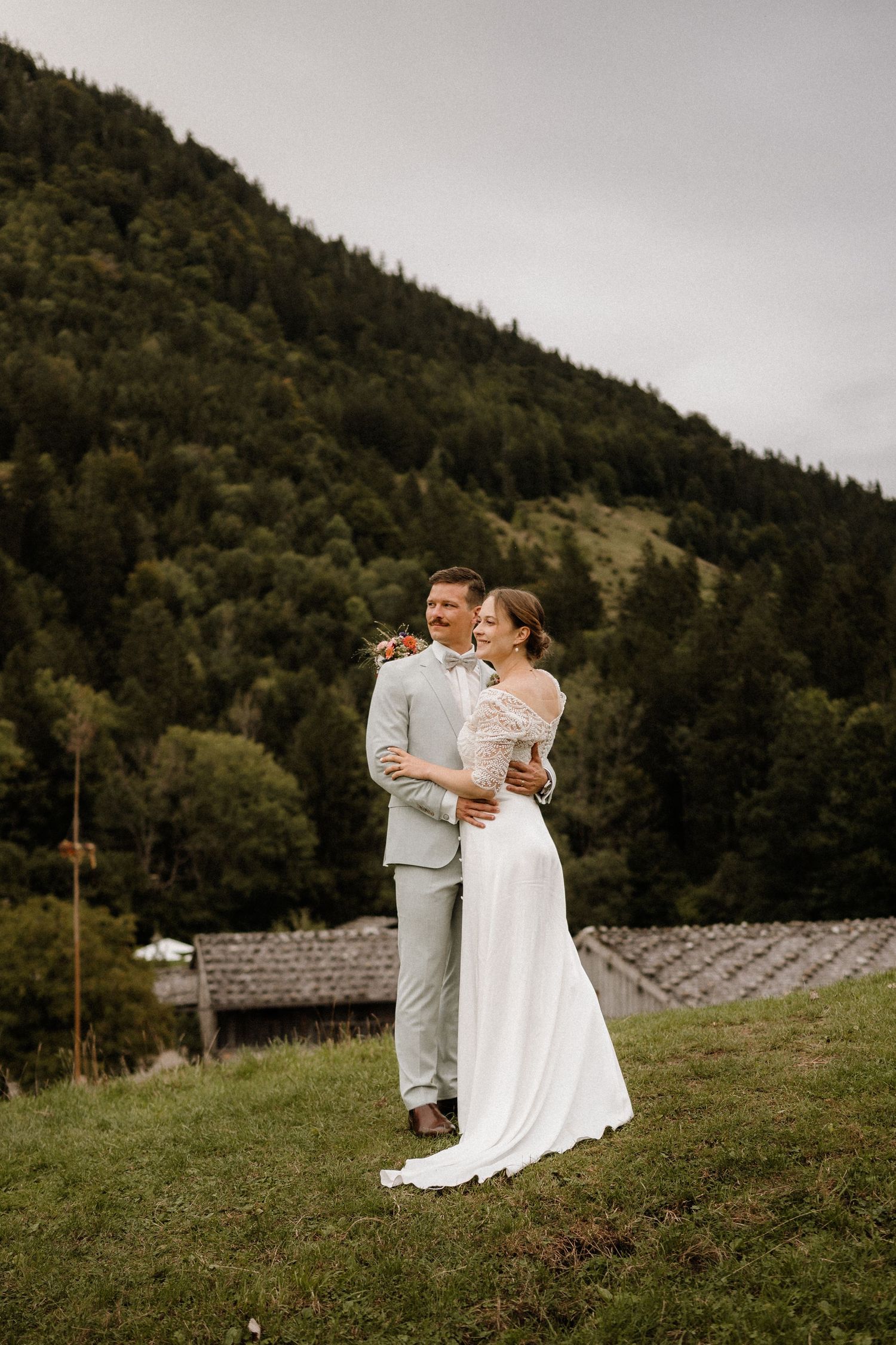 Newlyweds in elegant attire embracing on hillside with mountain backdrop.