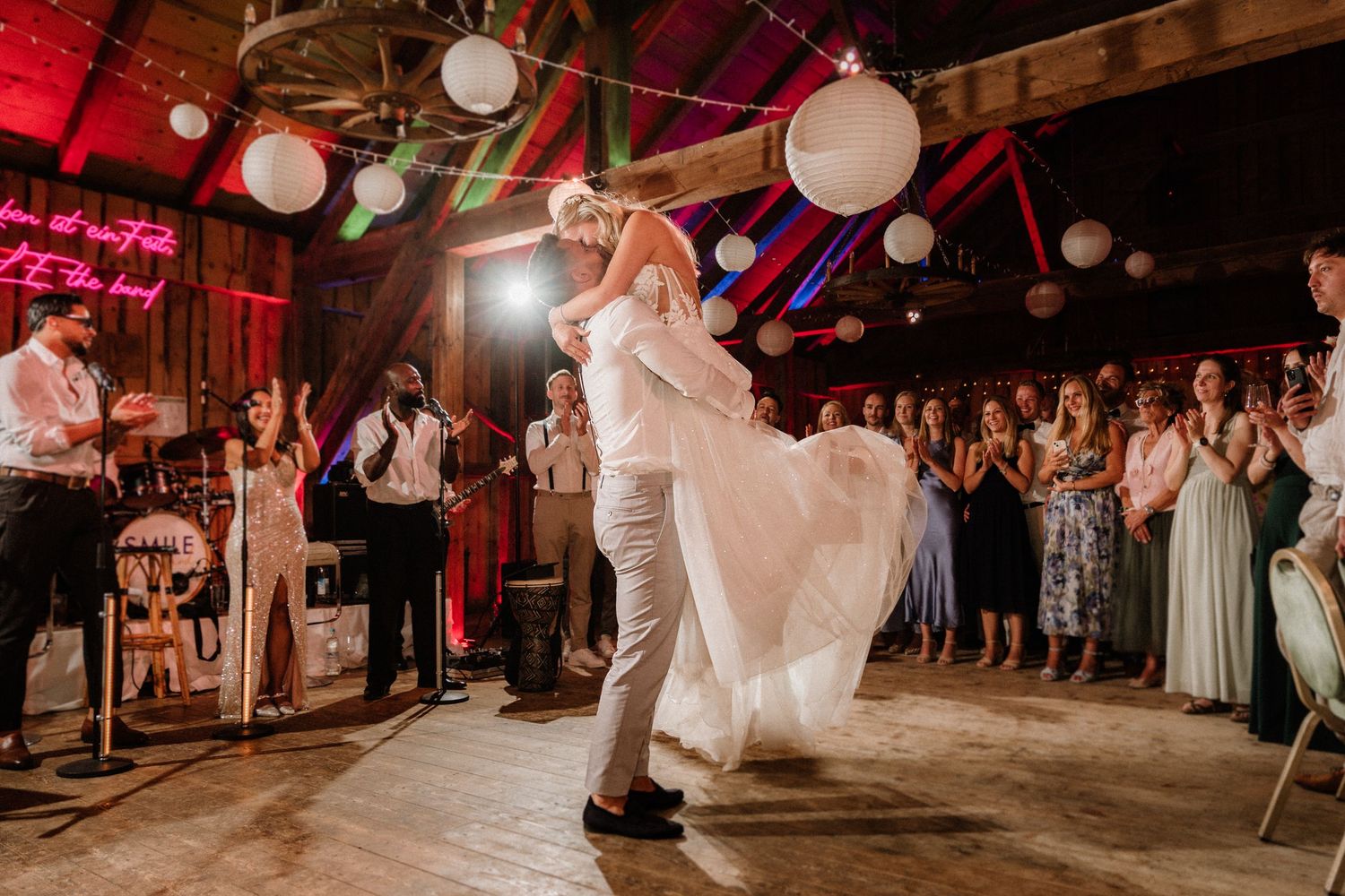 Couples dancing at a wedding reception in a barn with red mood lighting and decorative lanterns.