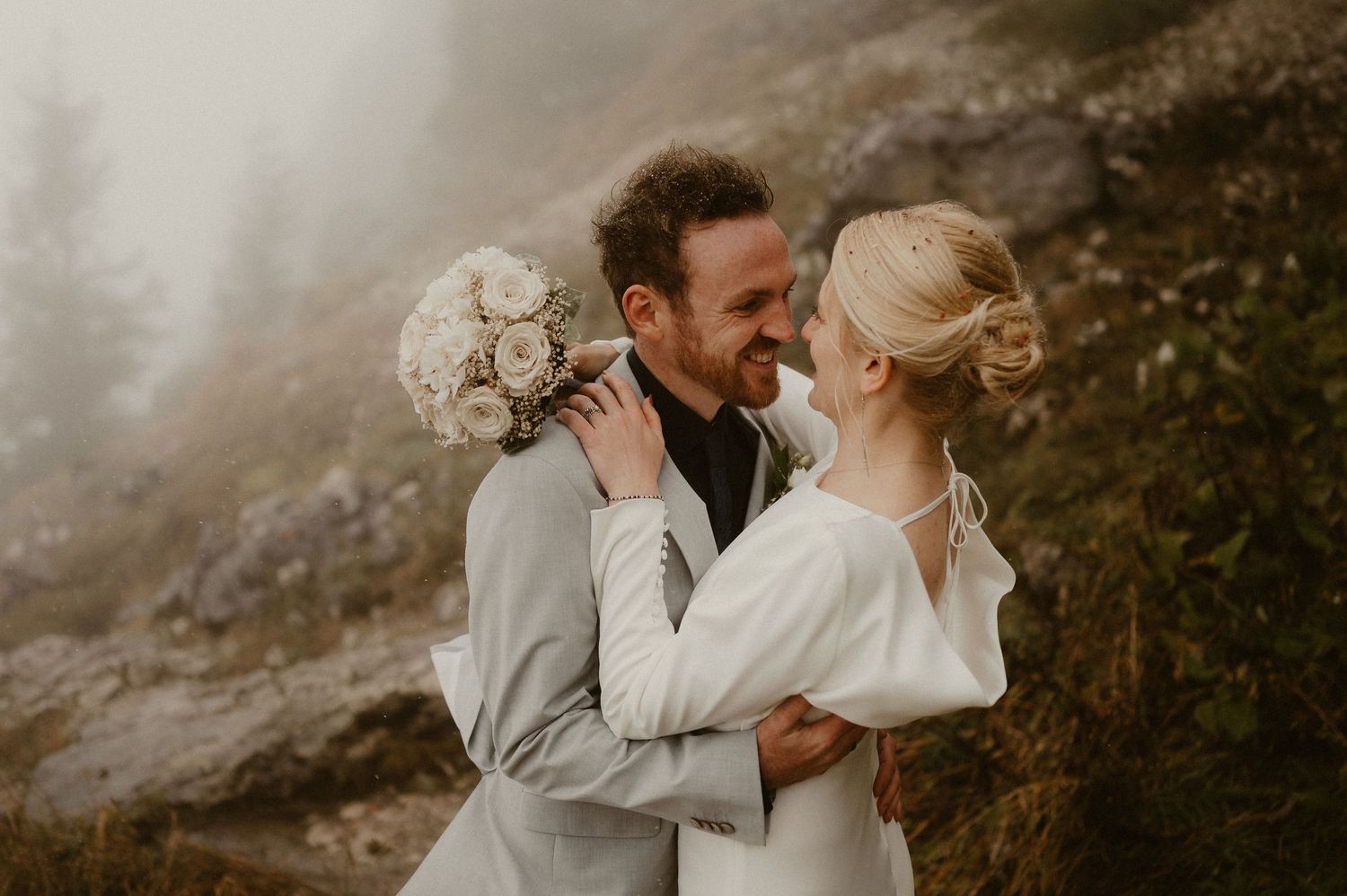 Romantic wedding couple embracing in misty mountain setting with white dress and grey suit.