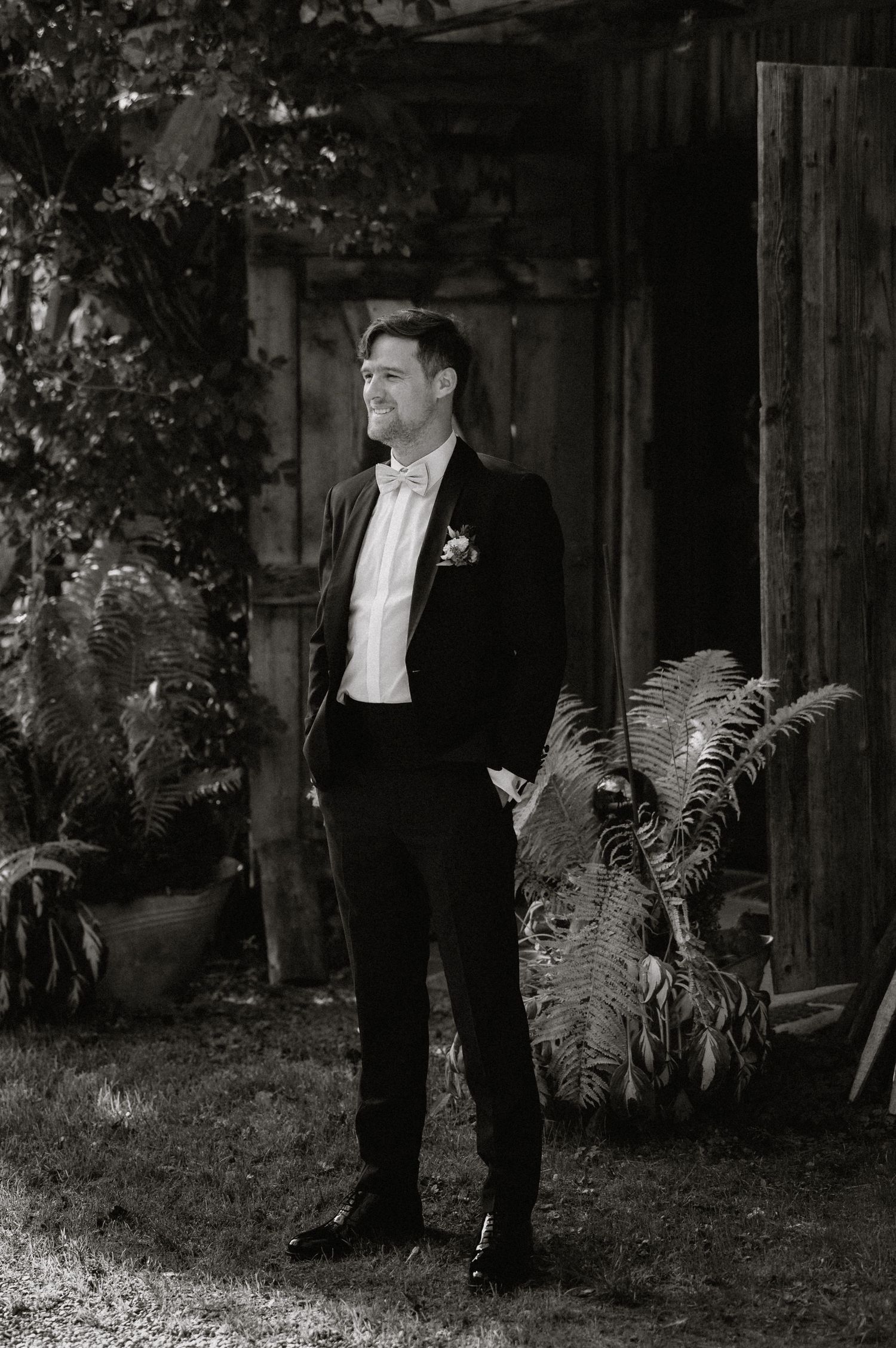 Groom in formal attire stands near rustic wooden structure in garden setting.