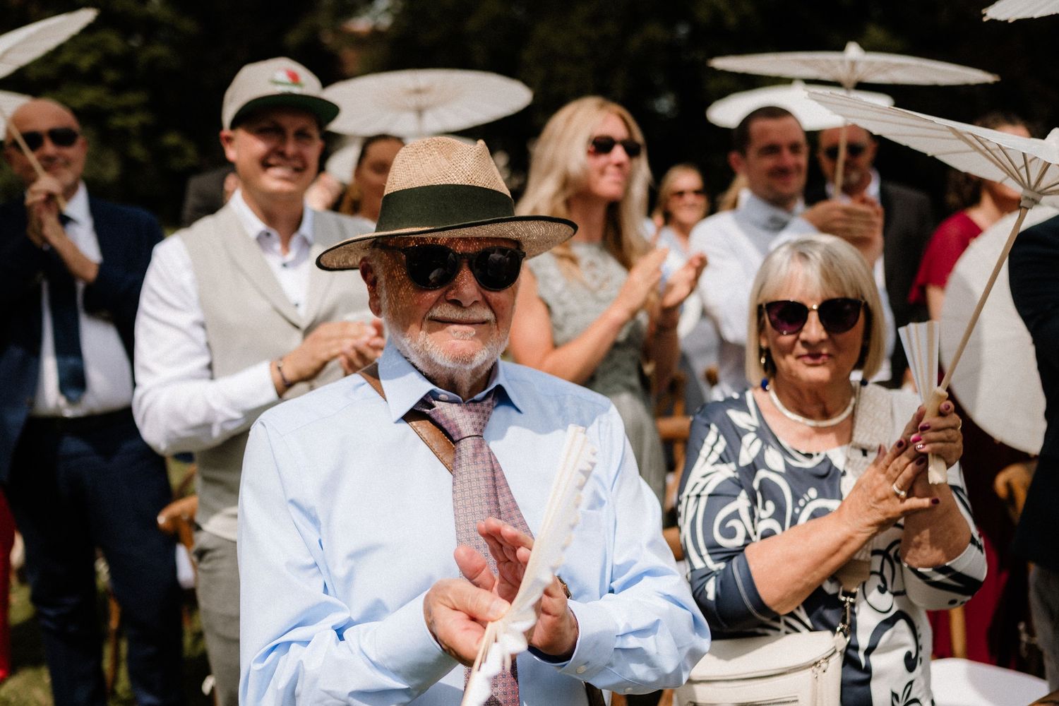 Wedding guests seated outdoors smile and watch the celebration unfold on a sunny day.