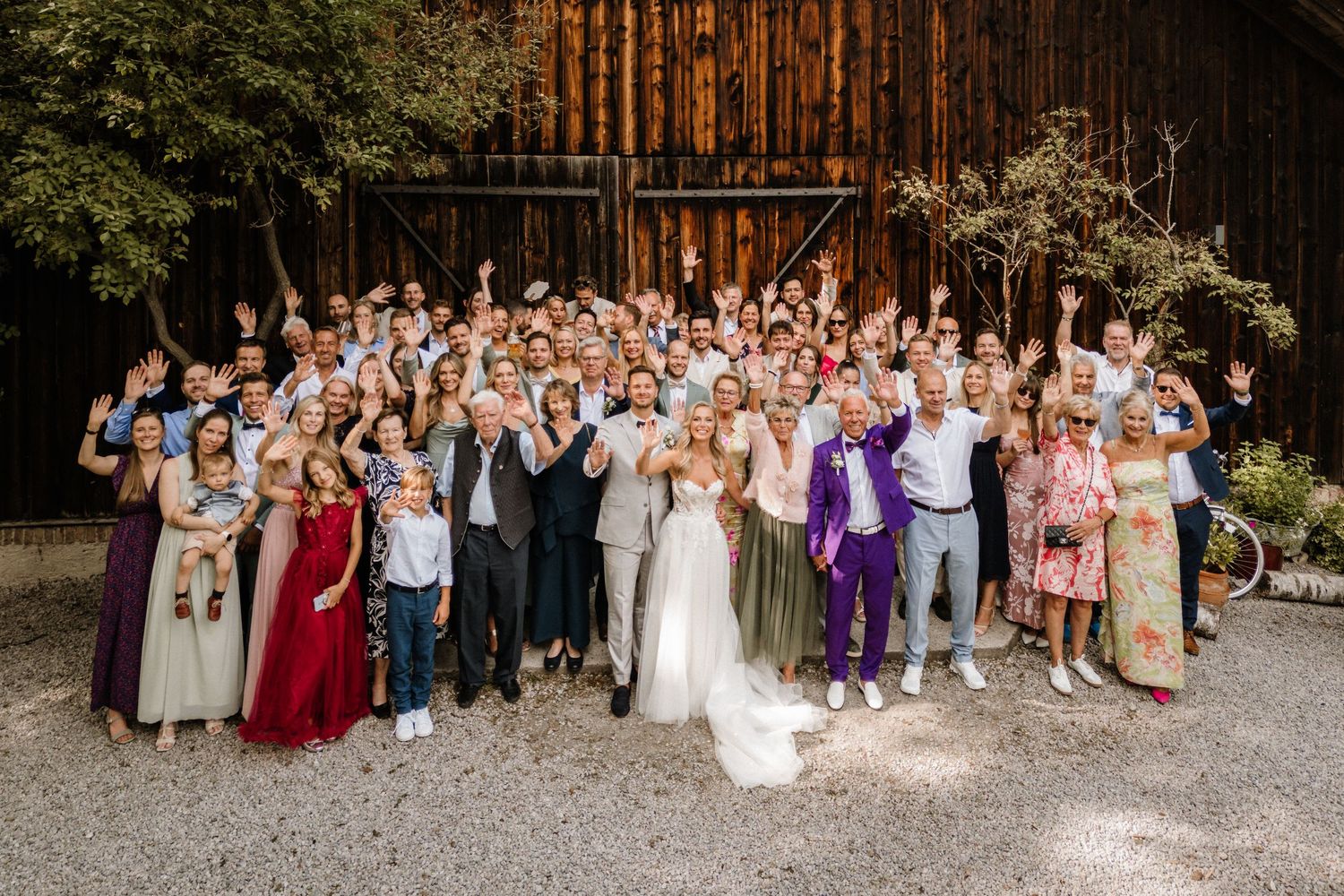 Large wedding party poses together outside rustic barn venue.
