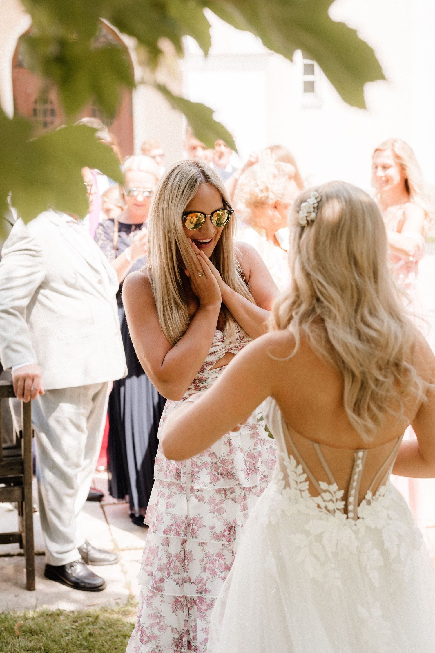 A surprised wedding guest reacts with joy while talking to the bride at an outdoor celebration.