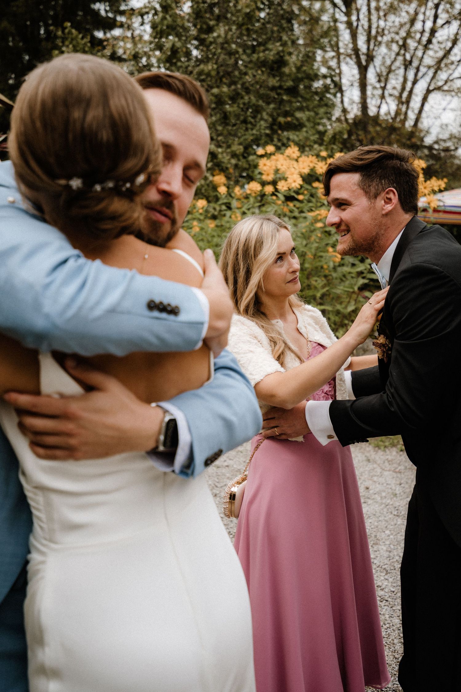 Wedding guests share warm embraces during an outdoor celebration with garden backdrop.