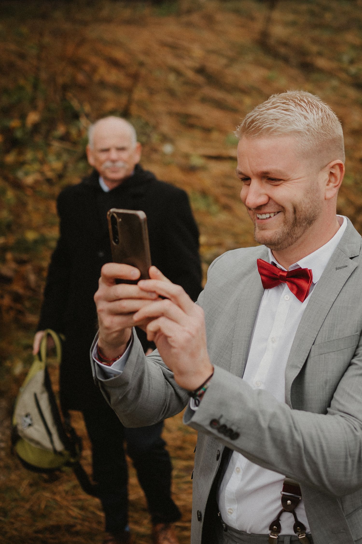 A groomsman in a grey suit and red bowtie smiles while looking at his phone during a wedding.